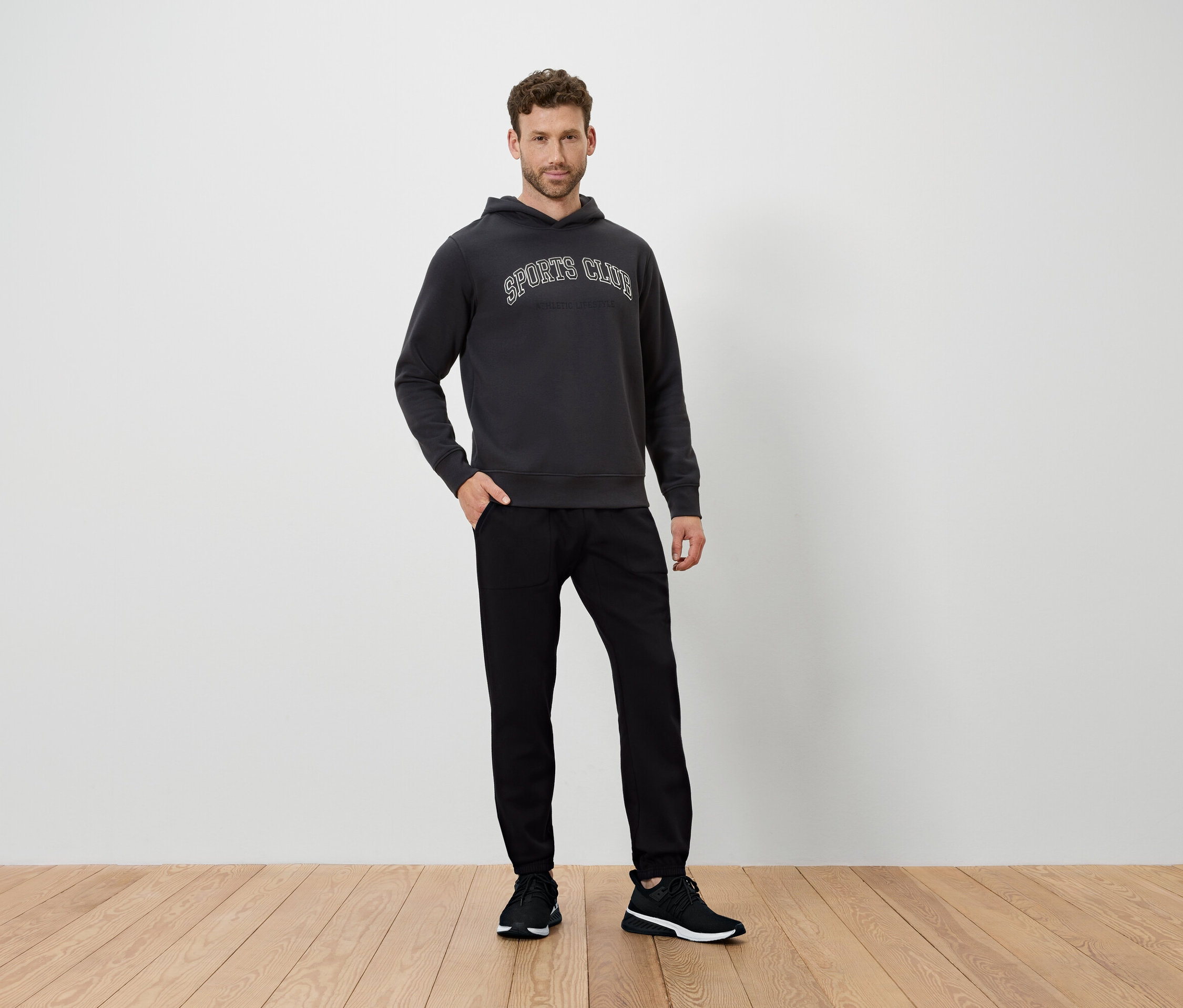 Mann steht auf Holzboden, trägt einen dunkelgrauen Hoodie und schwarze Hosen.