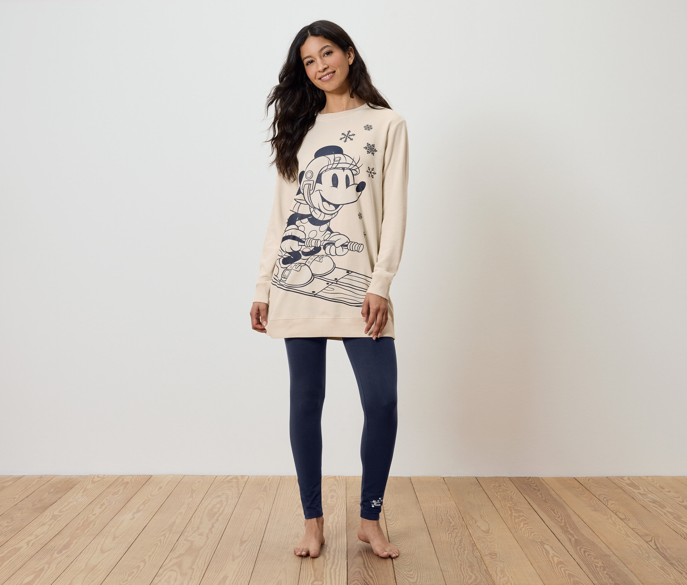 Frau steht barfuß und trägt ein langes beiges Sweatshirt mit Print und dunkelblaue Leggings.