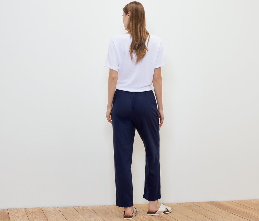 Eine Frau steht mit dem Rücken zur Kamera und trägt eine dunkelblaue Leinenhose, ein weißes T-Shirt und Sandalen.