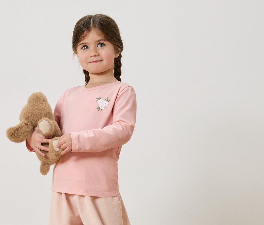 Mädchen mit Zöpfen hält einen Teddybären, trägt ein rosafarbenes Langarmshirt mit Blumenapplikation und rosafarbene Shorts.