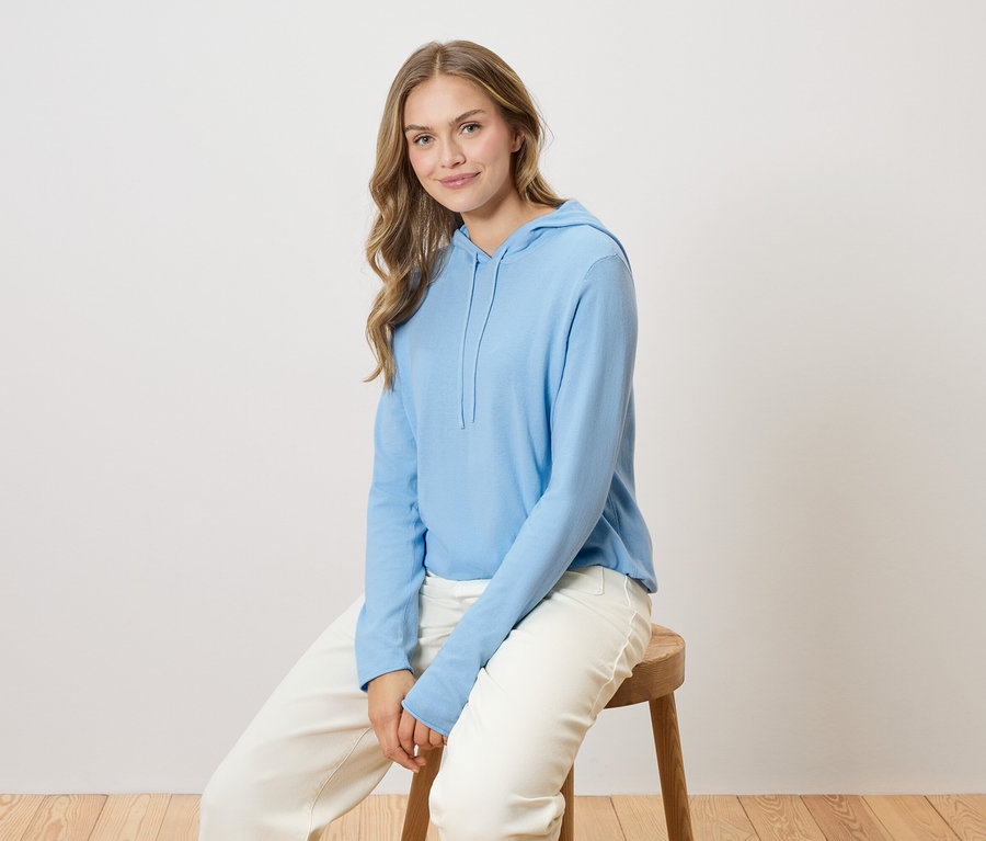 Frau mit langen blonden Haaren sitzt auf einem Holzstuhl und trägt einen hellblauen Kapuzenpullover und eine weiße Hose.