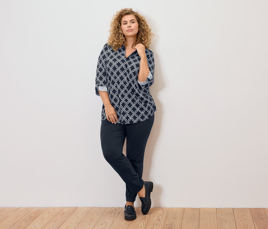 Frau mit lockigen blonden Haaren, bekleidet mit einer blau-weiß gemusterten Bluse, dunkler Hose und schwarzen Schuhen, lehnt an einer Wand.