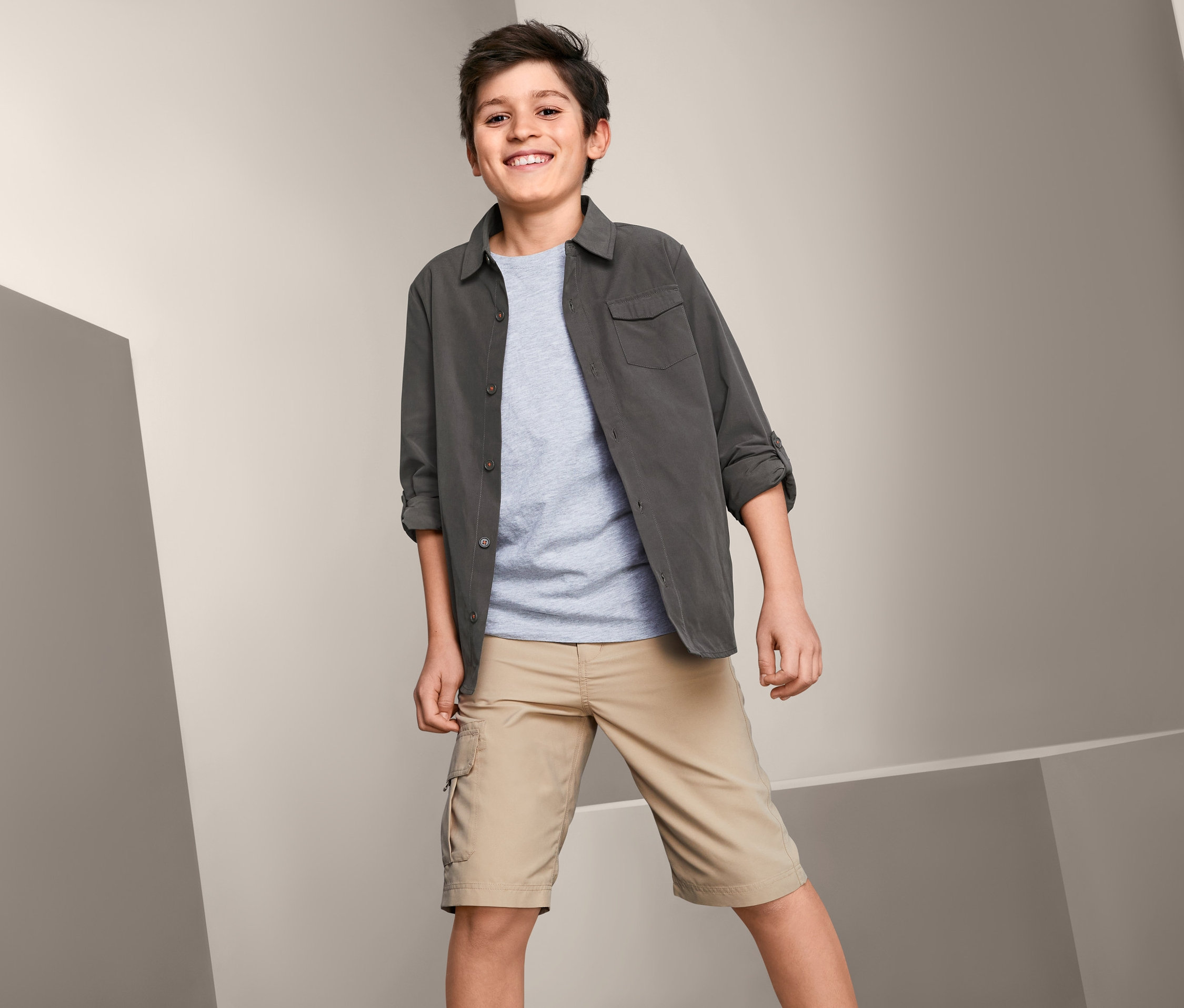 Ein lächelnder Junge posiert in einem grauen Funktions-Cargohemd und beigen Shorts.