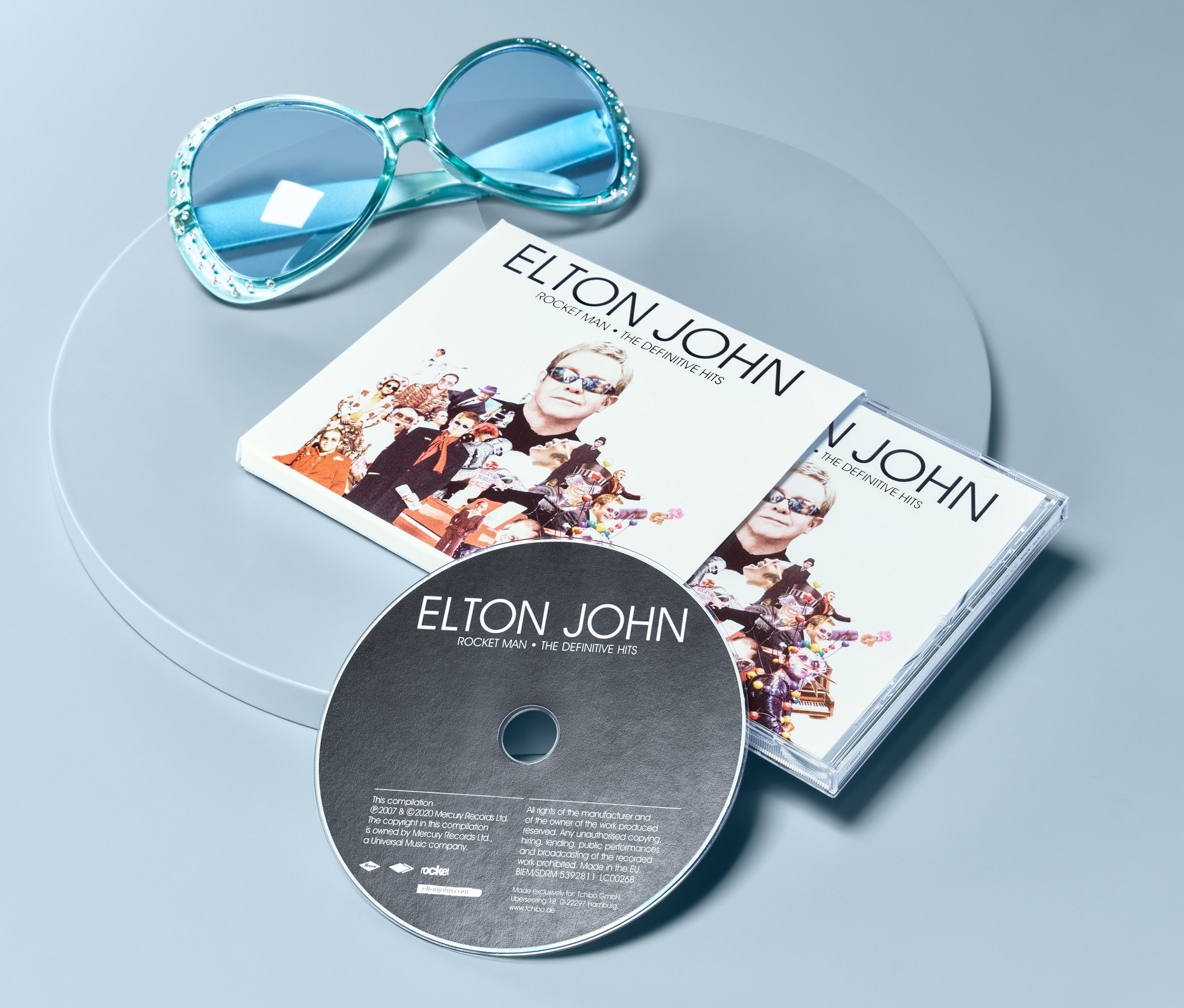 Blaue Sonnenbrille und CD Elton John – »Rocket Man: The Definitive Hits« auf grauem Untergrund.