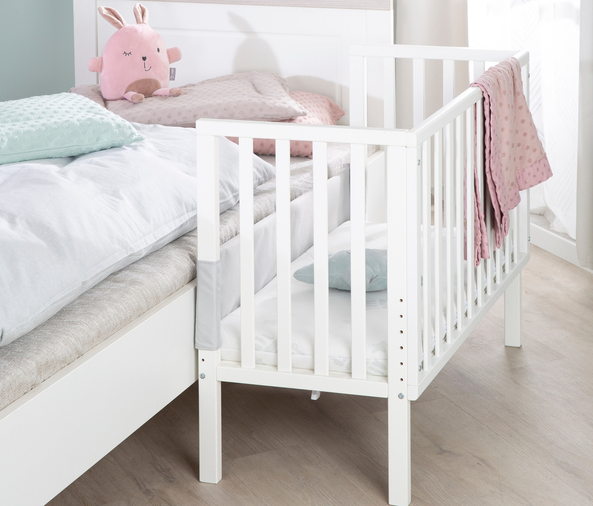 Ein weißes roba 2-in-1-Beistellbett »safe asleep®« steht neben einem großen Bett. Auf dem großen Bett liegt ein rosa Stoffhase.