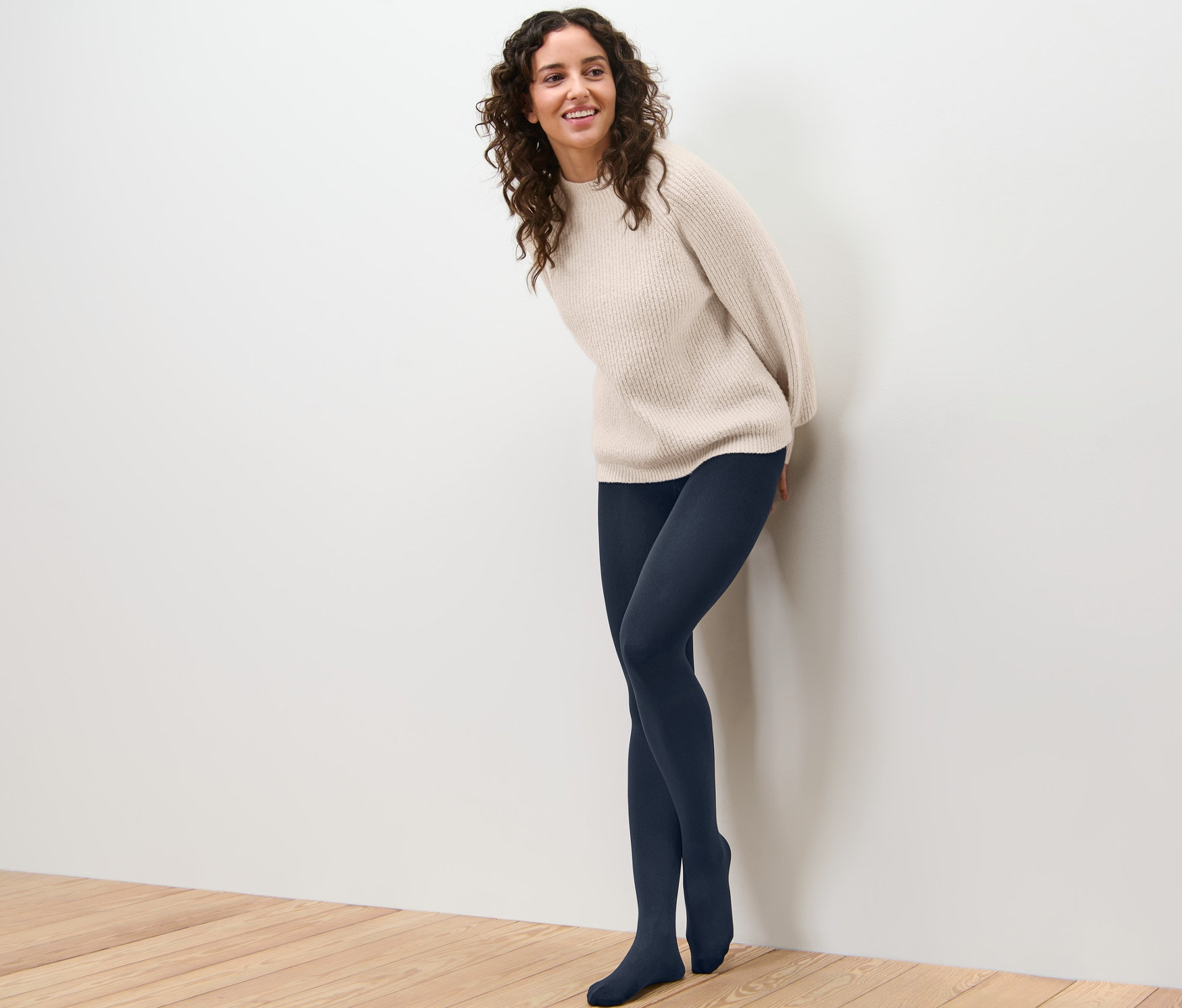 Lächelnde Frau, die sich an eine Wand lehnt, trägt eine Thermostrumpfhose und einen Pullover.