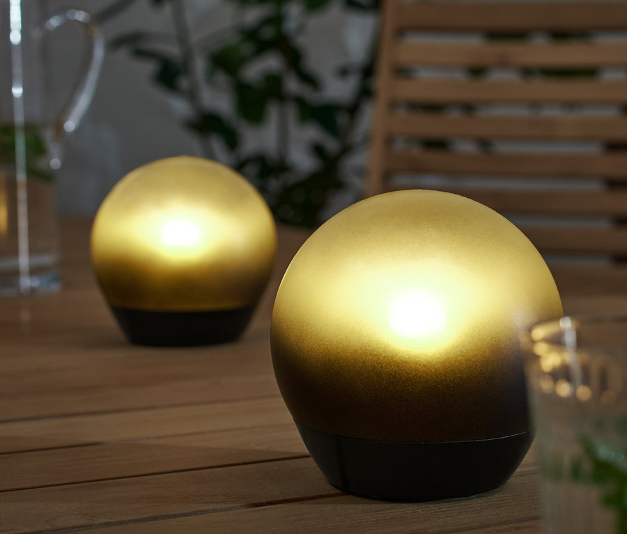 Zwei LED-Kugelleuchten stehen auf einem Holztisch, neben einem Glas und einem Krug mit Wasser.