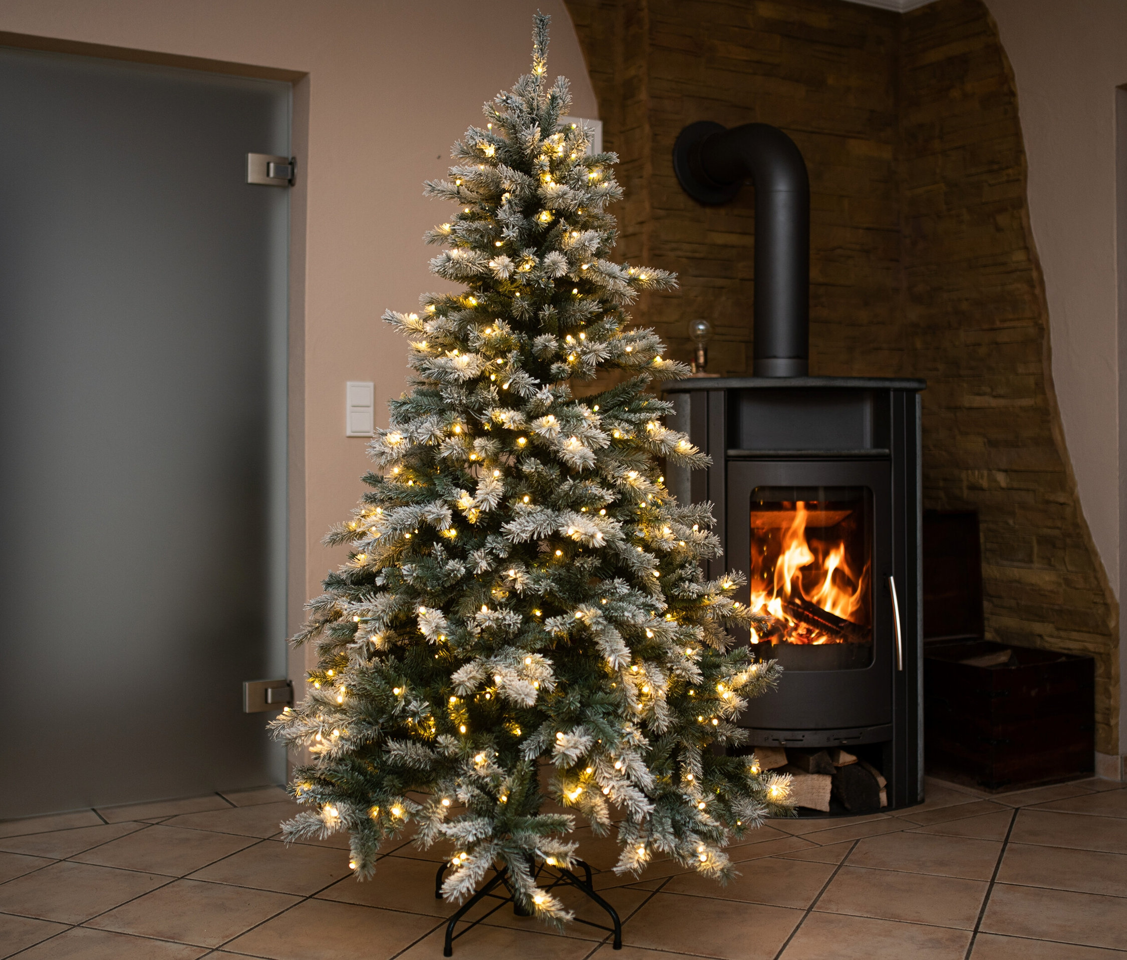 Beleuchteter, beschneiter künstlicher Weihnachtsbaum »Fichte« steht neben einem schwarzen Kaminofen.