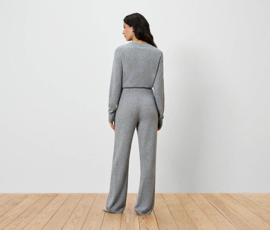 Frau steht mit dem Rücken zur Kamera und trägt ein graues Strickoberteil mit langen Ärmeln und eine weite Hose.