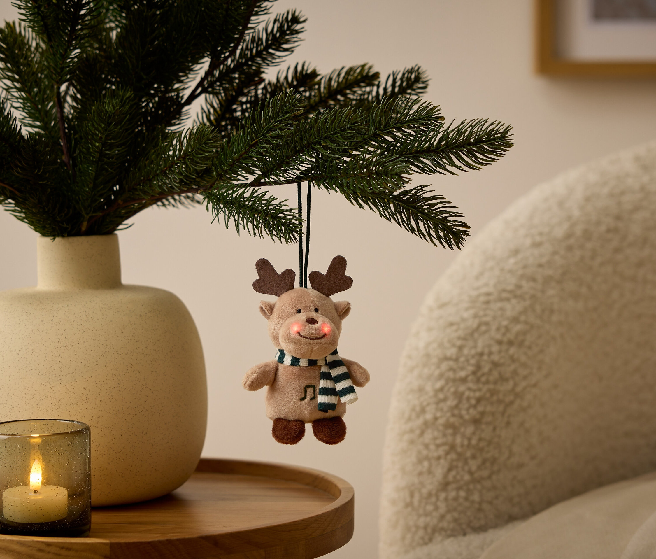 Ein Rentier-Ornament hängt an einem Zweig in einer Vase neben einer Kerze auf einem Tisch.