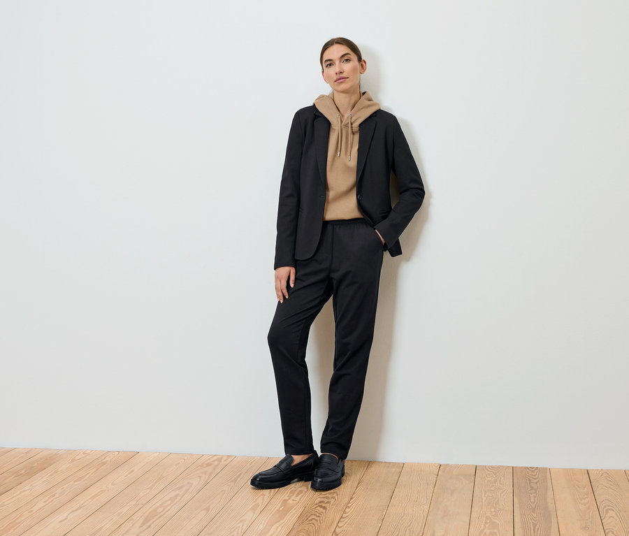 Eine Frau posiert und trägt einen schwarzen Blazer, einen braunen Kapuzenpullover und eine schwarze Hose.