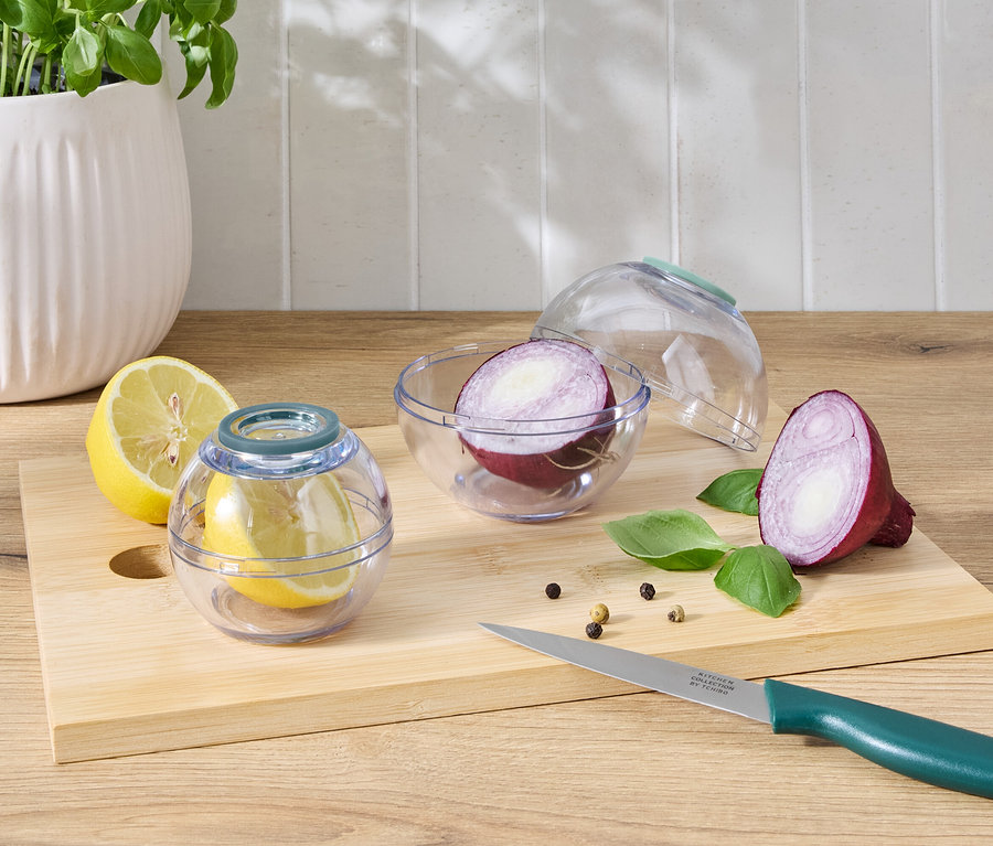 Eine Zitronenhälfte in einem transparenten Behälter neben einer Zitronenhälfte und einer halben roten Zwiebel in einem transparenten Behälter neben einer halben roten Zwiebel auf einem Schneidebrett mit Messer.