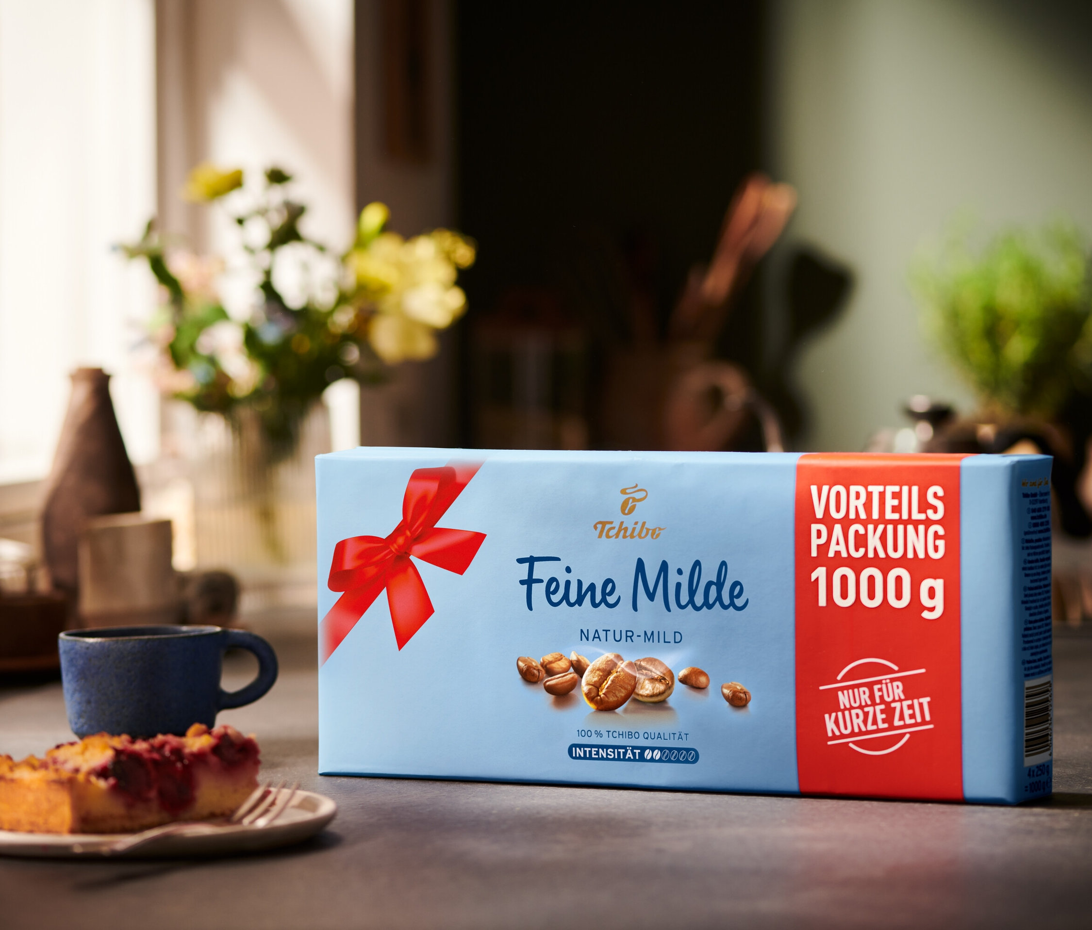 Eine Packung Feine Milde Kaffee steht neben einer Tasse und einem Stück Kuchen auf einem Tisch.