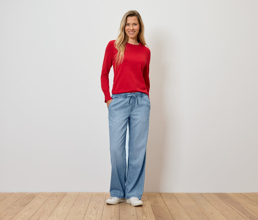 Eine Frau in einem roten Pullover und hellblauen Schlaghosen steht auf einem Holzboden.