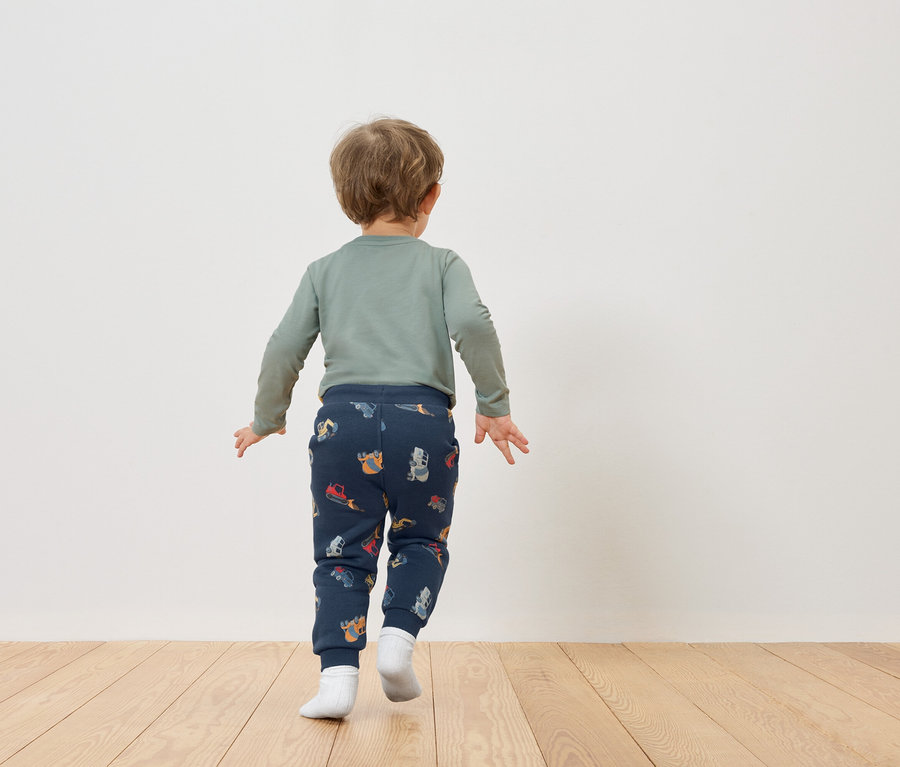Ein Kleinkind mit blonden Haaren, von hinten gesehen, trägt ein grünes Langarmshirt und dunkelblaue Hosen mit Fahrzeugmuster.