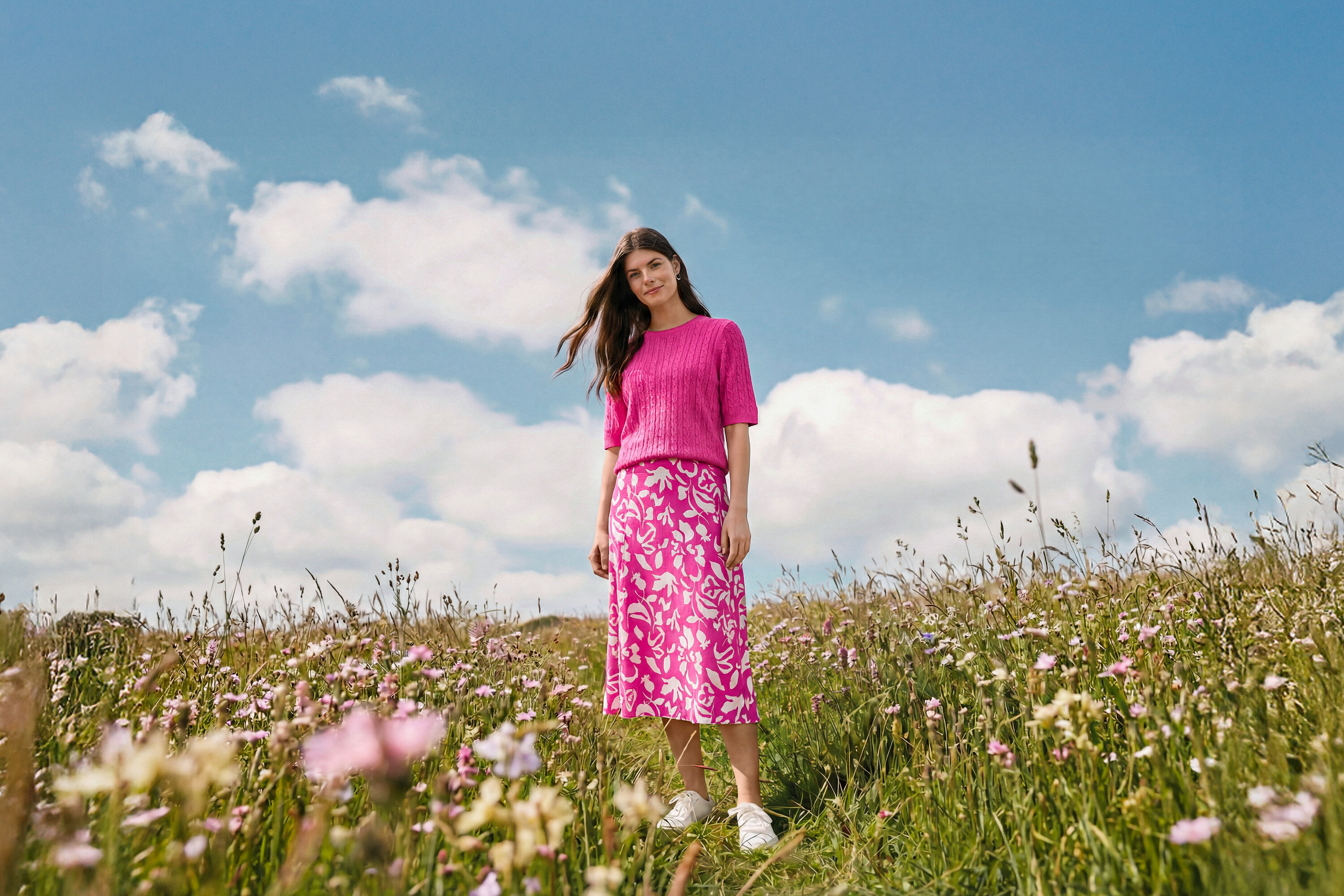 Eine Frau steht auf einer Wiese und trägt einen bedruckten Midi-Rock, Creolen Wave und Leder-Sneaker.