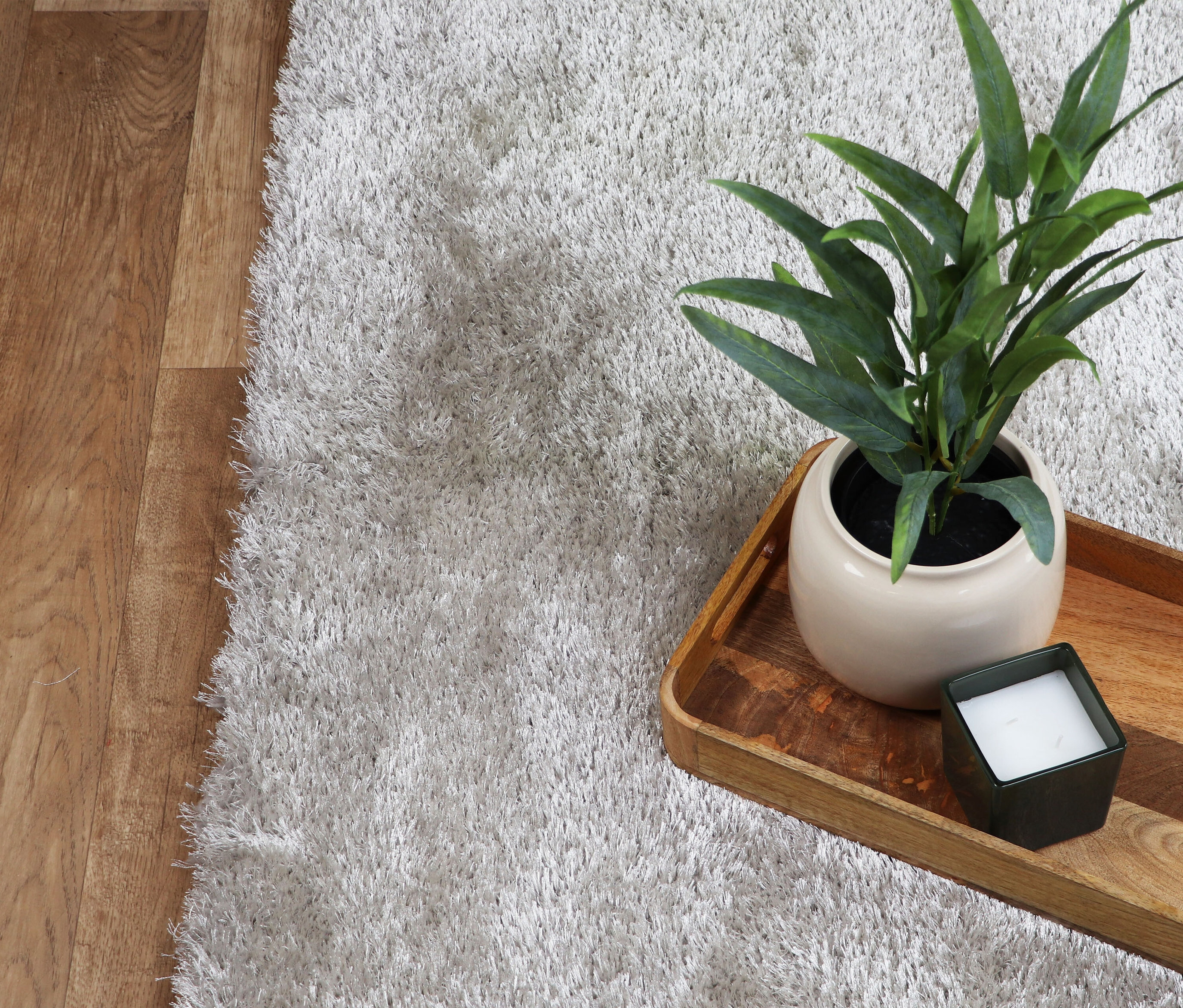 Nahaufnahme eines hellgrauen Teppichs »Silky« neben einem Holzboden. Auf dem Teppich steht ein Tablett mit einer Pflanze und einer Kerze.