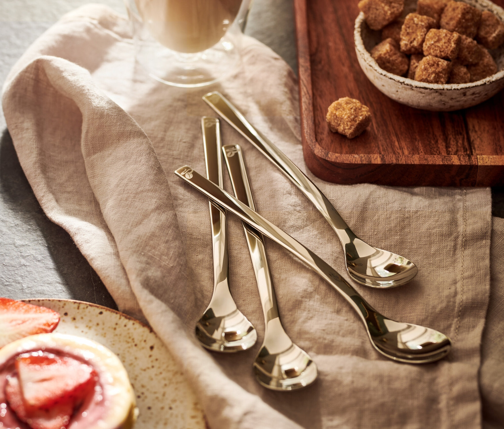 Glas Latte Macchiato, daneben eine Schale mit braunem Zucker und ein Törtchen mit Erdbeeren. Auf der Serviette liegt ein 4er Set Latte Macchiato Löffel.
