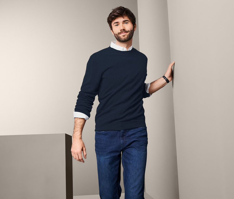 Mann lehnt an Wand, trägt Cashmere-Pullover mit Rundhalsausschnitt, Jeans und Uhr.