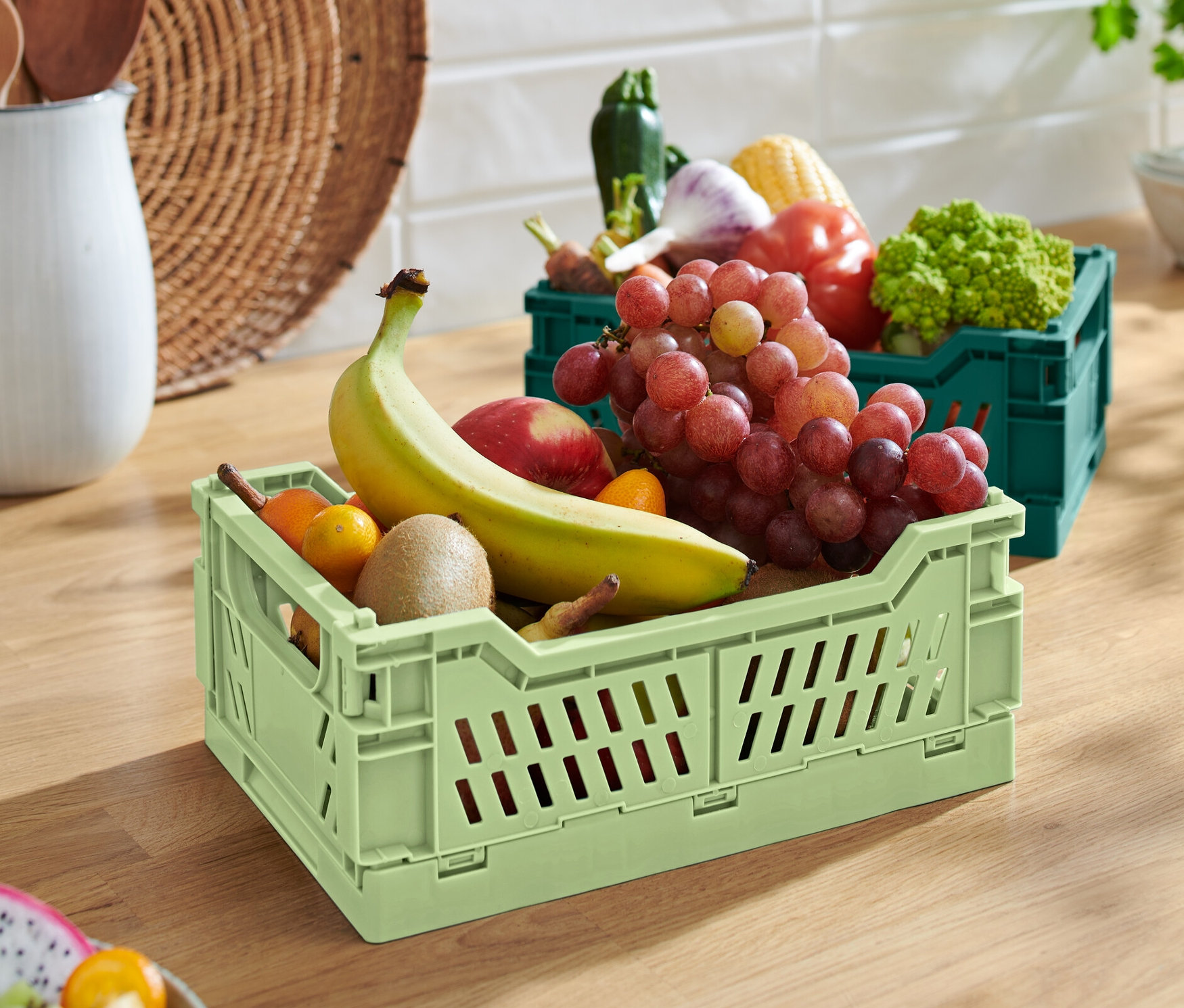 Eine grüne klappbare Ordnungskiste mit Banane, Apfel, Kiwi und Trauben. Im Hintergrund eine grüne klappbare Ordnungskiste mit Gemüse.