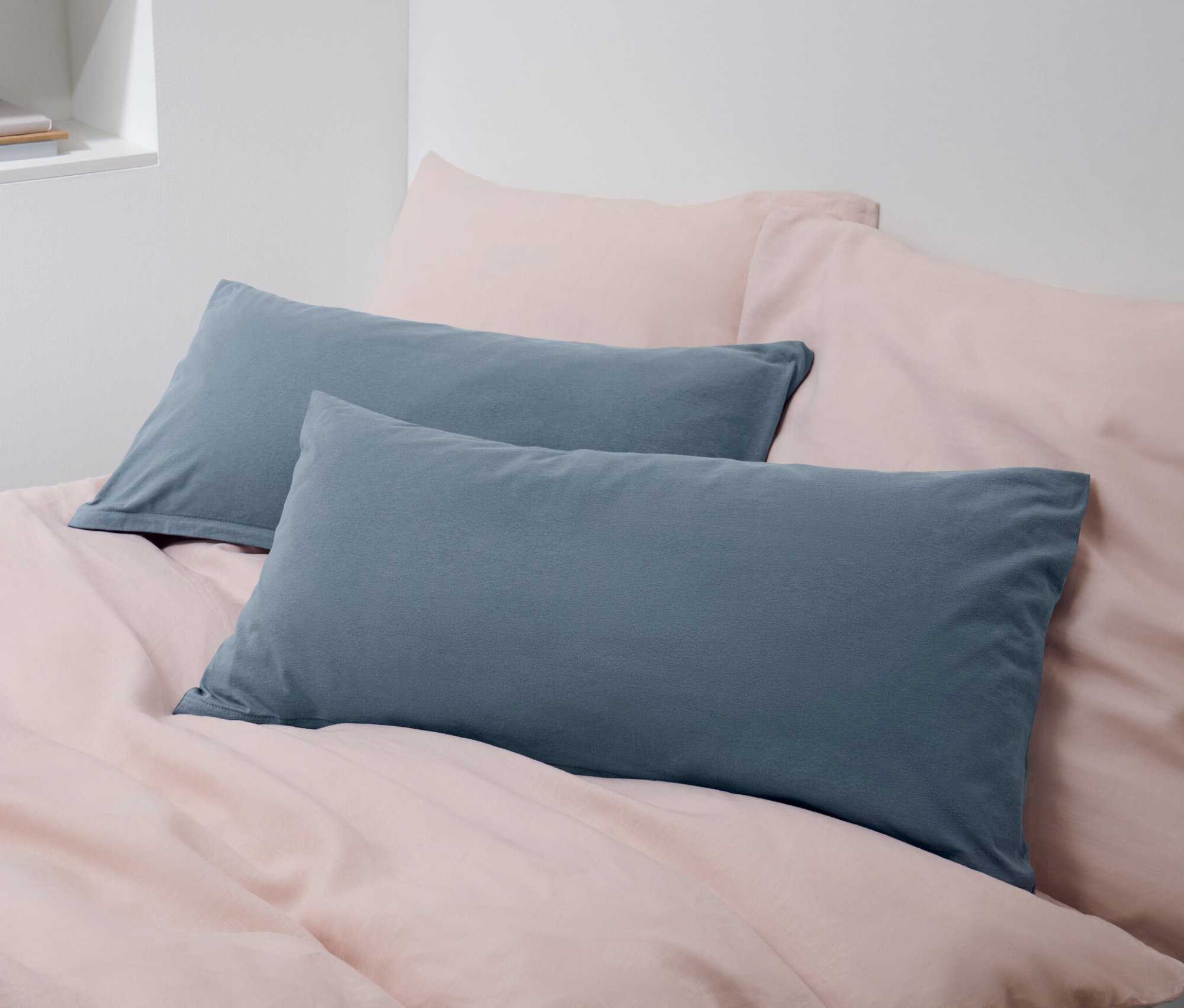 Zwei blaue Jersey-Kopfkissenbezüge liegen auf einem Bett mit rosa Bettwäsche.
