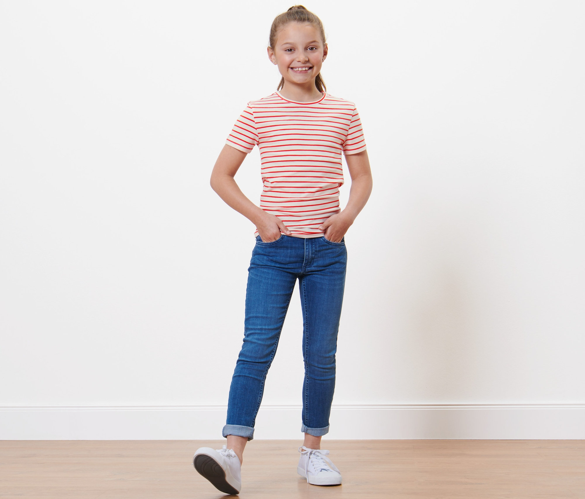 Ein Mädchen mit einem rot gestreiften T-Shirt und Jeans steht mit den Händen in den Taschen. 3 Kinder-T-Shirts.