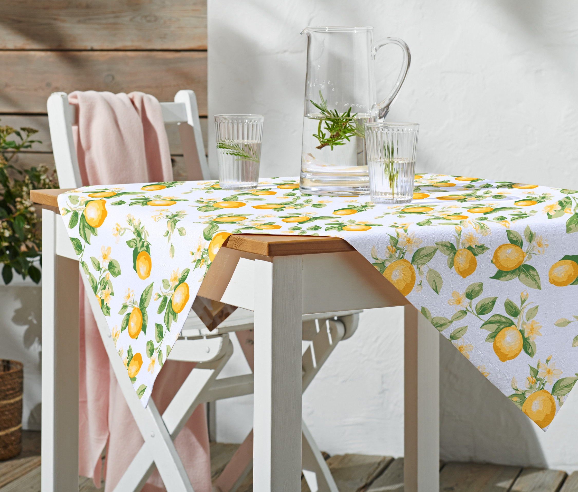 Ein Tisch mit einer Baumwoll-Tischdecke mit Zitronenmuster, Gläsern und einem Krug Wasser steht draußen.