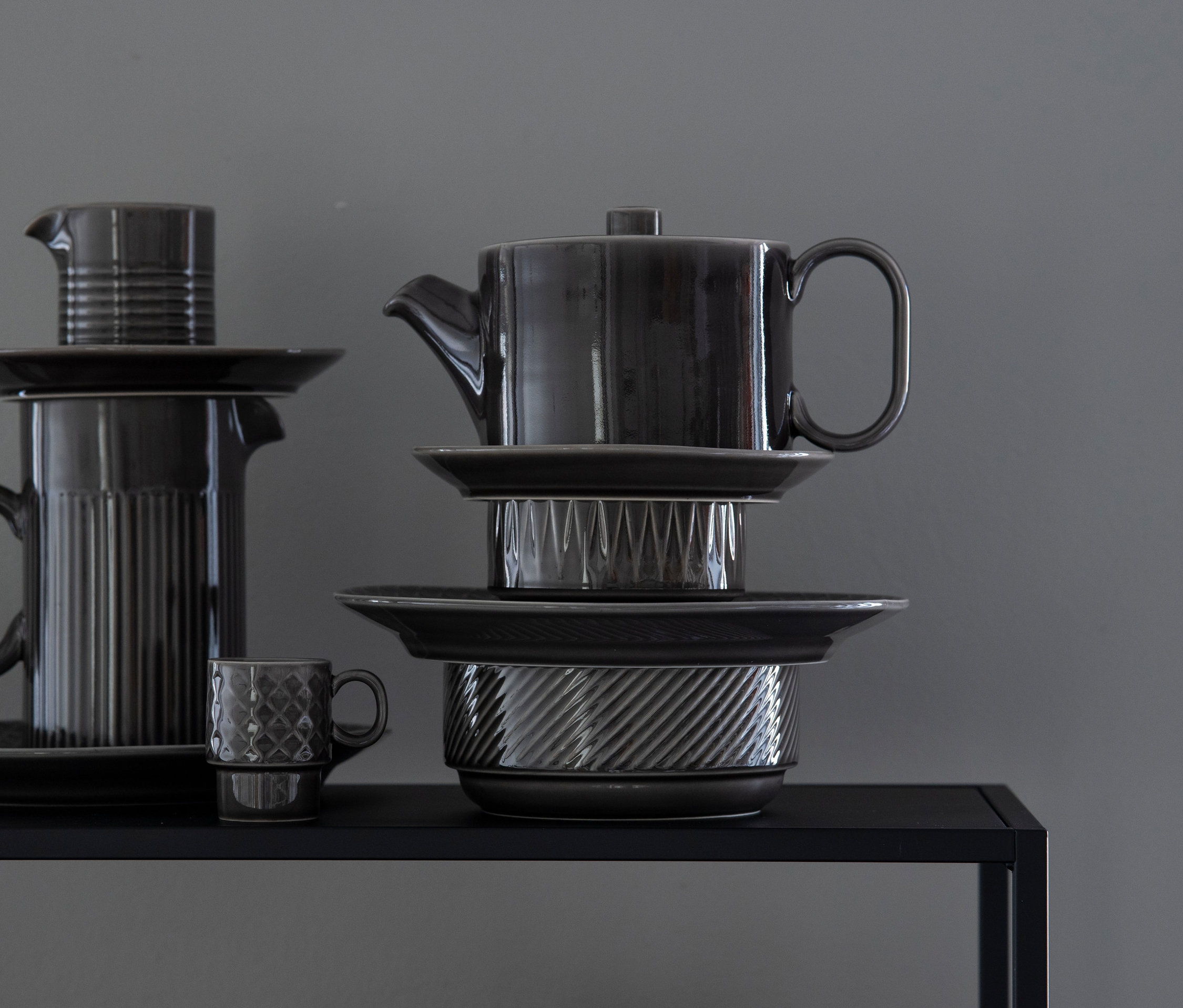 Graue Keramik Teekanne, Krug, Milchkännchen, tiefer Teller und Teller sind auf einem schwarzen Regal gestapelt.