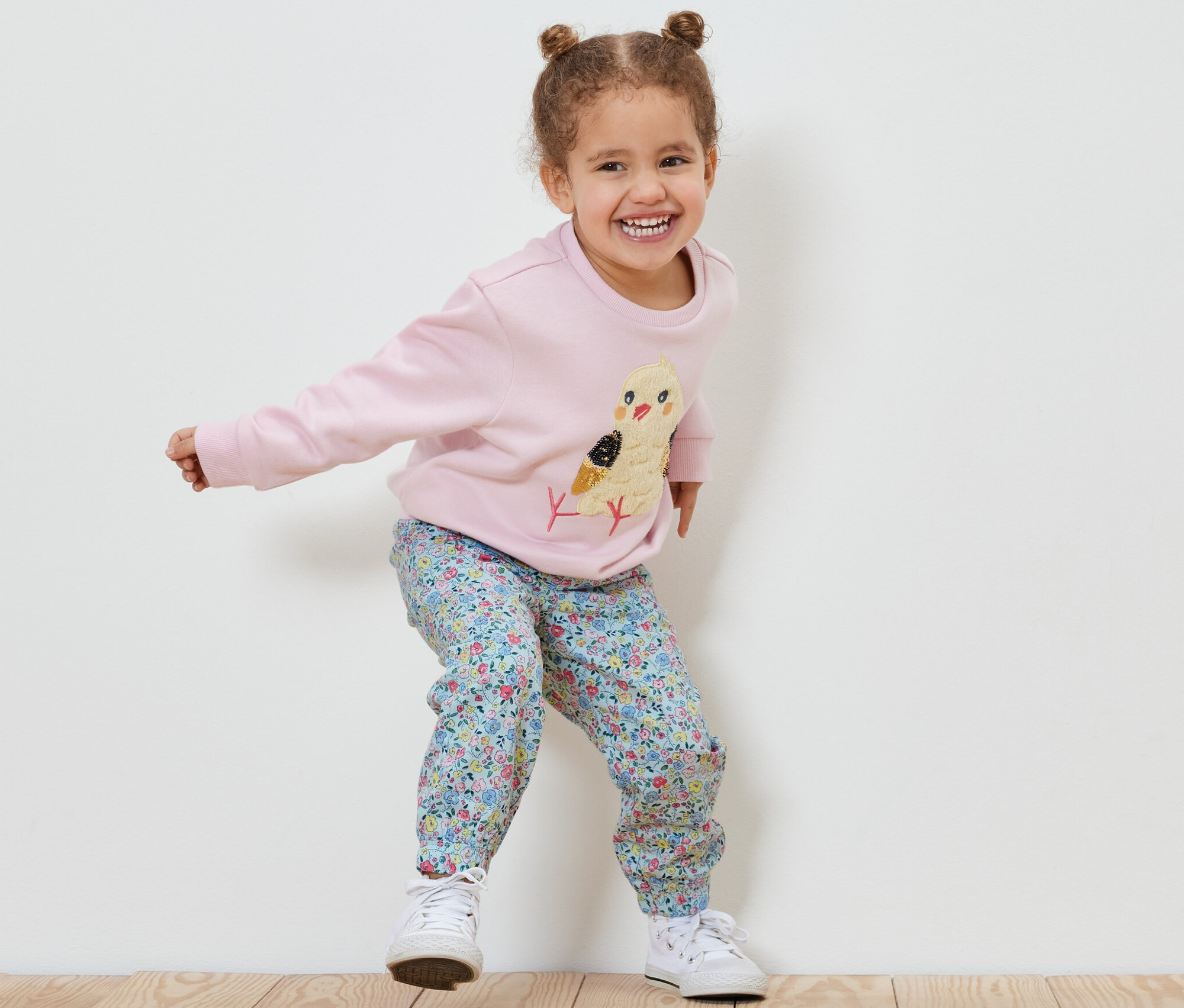 Lächelndes kleines Mädchen springt und trägt einen rosa Pullover mit Küken und Kinder-Pull-on-Pants – Fit »Ella«.