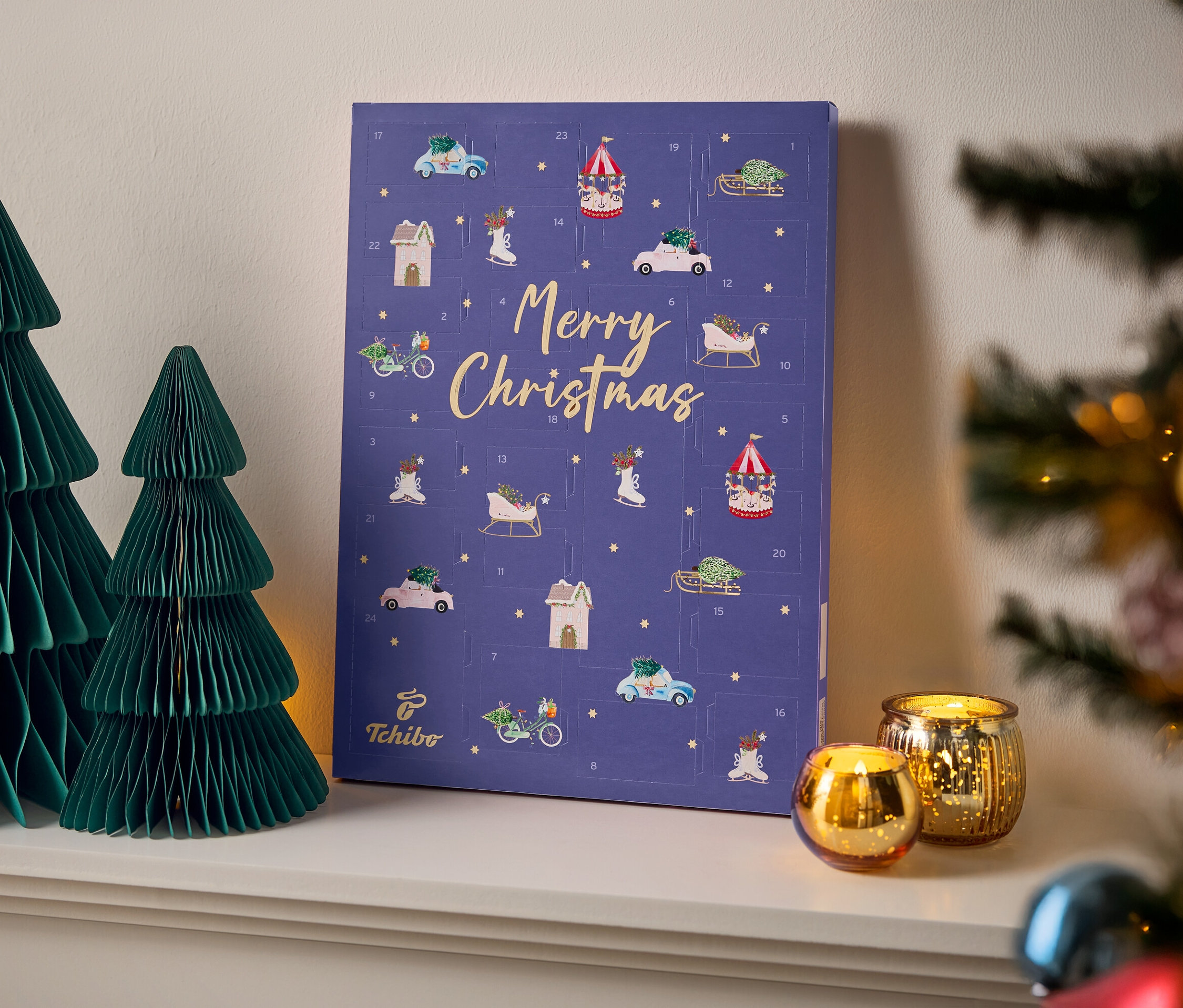 Auf einem weißen Regal stehen zwei grüne Papierbäume neben einem violetten Tchibo Adventskalender mit der Aufschrift Frohe Weihnachten und zwei goldenen Kerzen.