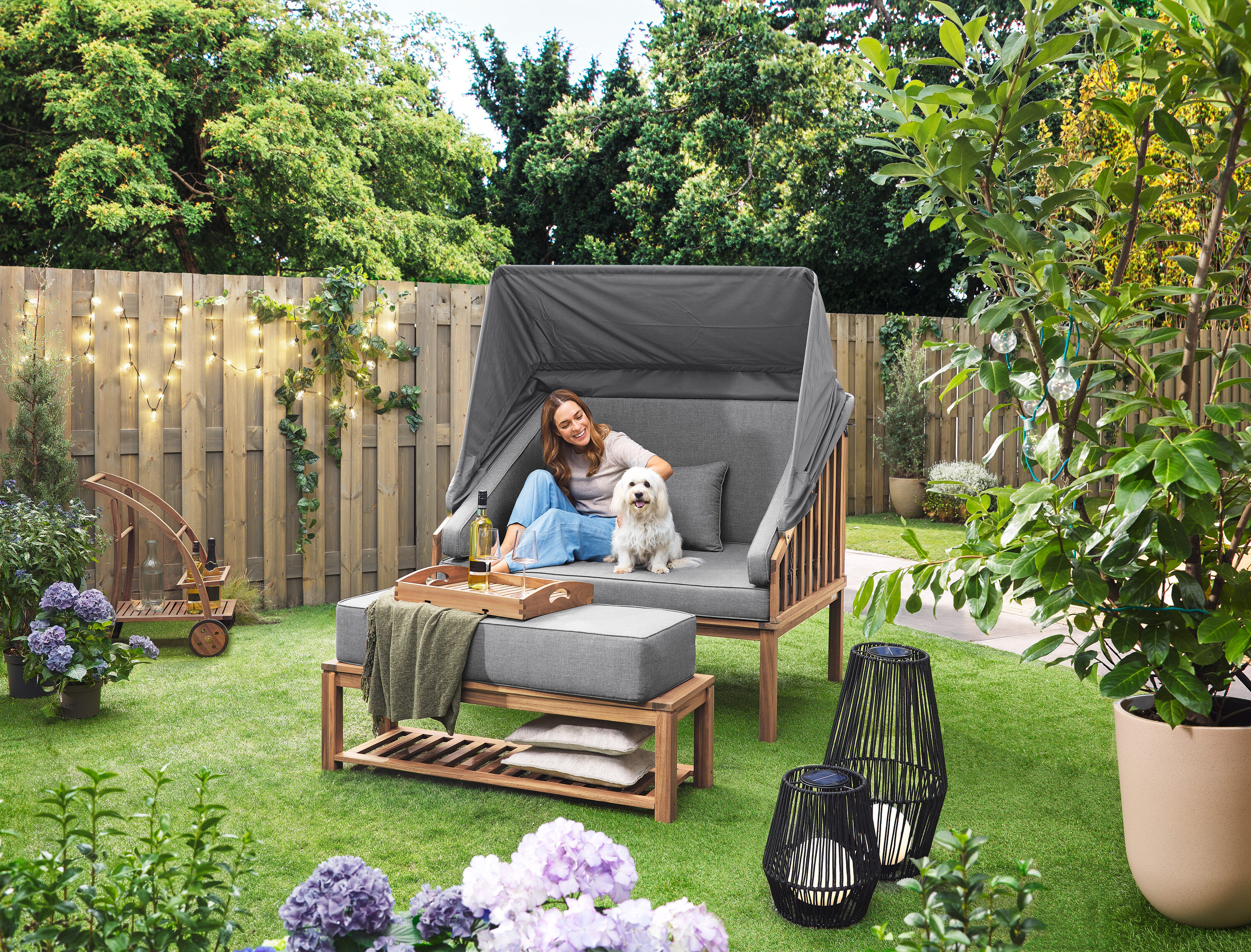 Eine Frau sitzt mit einem Hund in einem Premium Lounge-Strandkorb mit Sonnenschutz-Dach. Eine Solar-Laterne mit Textilband, groß und eine Solar-Laterne mit Textilband, klein stehen auf dem Rasen. Ein Barwagen mit abnehmbarem Tablett steht im Hintergrund.