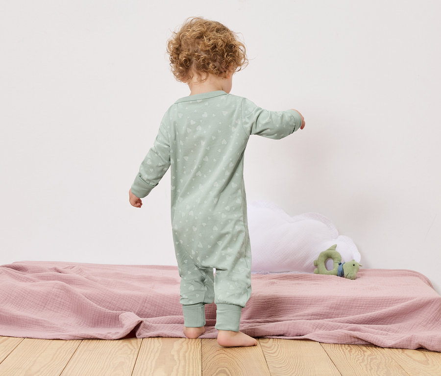 Ein Kleinkind steht mit dem Rücken zur Kamera auf einem Bett mit einer rosa Decke und einem grünen Spielzeug.