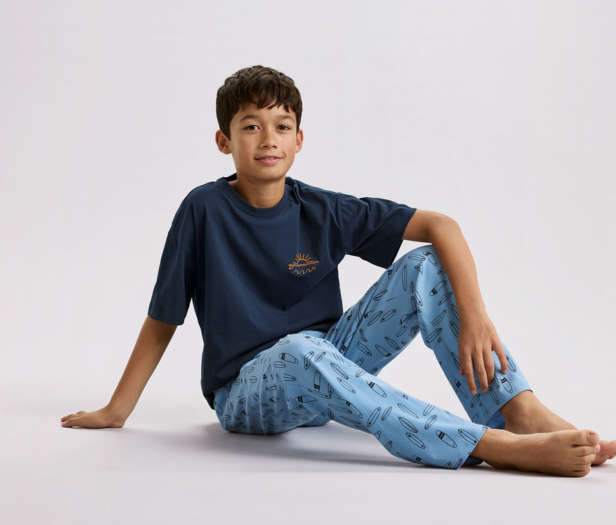 Ein Junge sitzt auf dem Boden, trägt ein marineblaues T-Shirt mit einer kleinen Sonne und Wellen darauf und hellblaue Pyjamahosen mit Surfbrettmuster.