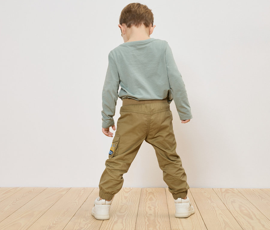 Ein Kind steht mit dem Rücken zur Kamera, trägt ein grünes Langarmshirt, khaki Hosen und weiße Turnschuhe.