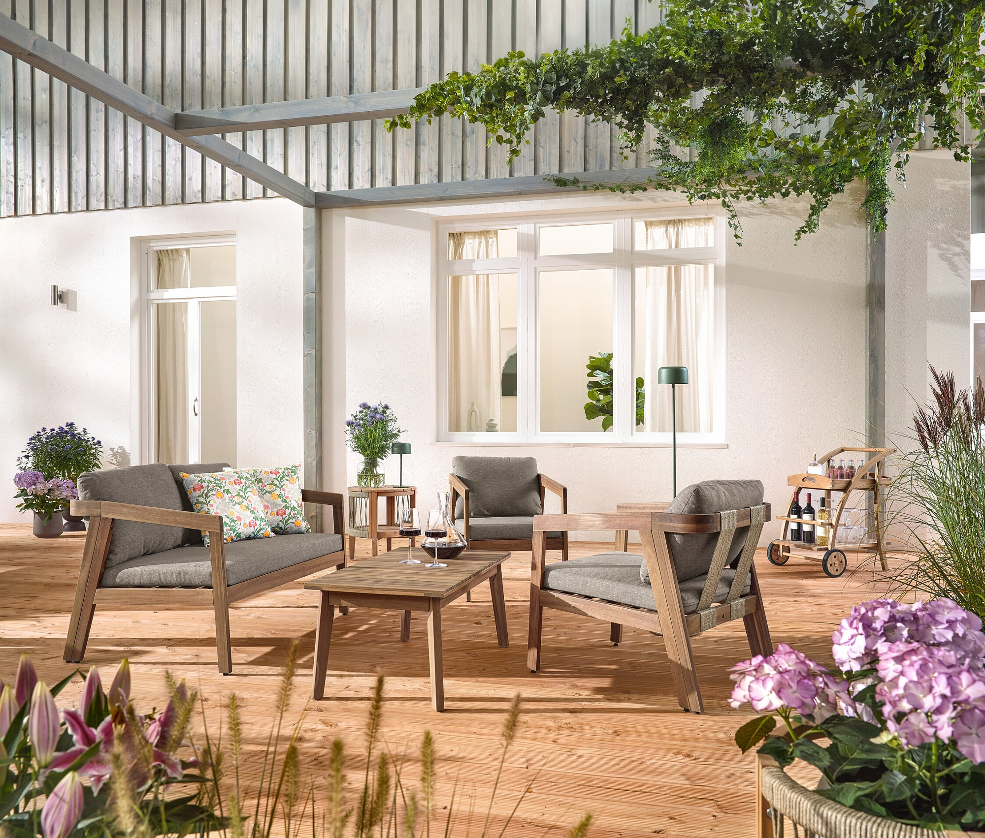 Auf einer Holzterrasse steht ein Garten-Lounge-Set mit Textilgeflecht, 4-tlg. mit 2 Outdoor-Kissen mit floralem Muster, ein Barwagen mit abnehmbarem Tablett und ein Garten-Beistelltisch mit abnehmbarer Tischplatte, Ø ca. 50 cm. Eine Solar-Akku-Tischleuchte, dschungelgrün und eine Solar-Akku-Stehleuchte, dschungelgrün sind auch zu sehen.