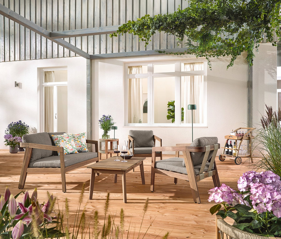 Auf einer Holzterrasse steht ein Garten-Lounge-Set mit Textilgeflecht, 4-tlg. mit 2 Outdoor-Kissen mit floralem Muster, ein Barwagen mit abnehmbarem Tablett und ein Garten-Beistelltisch mit abnehmbarer Tischplatte, Ø ca. 50 cm. Eine Solar-Akku-Tischleuchte, dschungelgrün und eine Solar-Akku-Stehleuchte, dschungelgrün sind auch zu sehen.