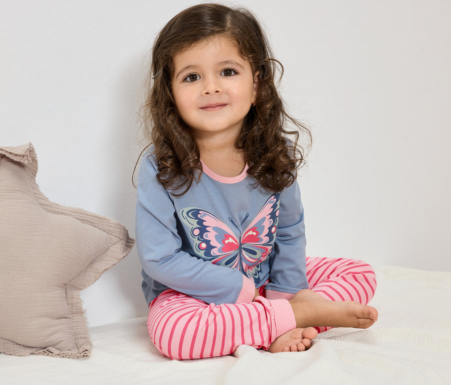 Ein junges Mädchen mit lockigem Haar sitzt auf einem Bett und trägt einen blauen Schlafanzug mit einem Schmetterlingsmotiv und rosa-gestreiften Hosen.