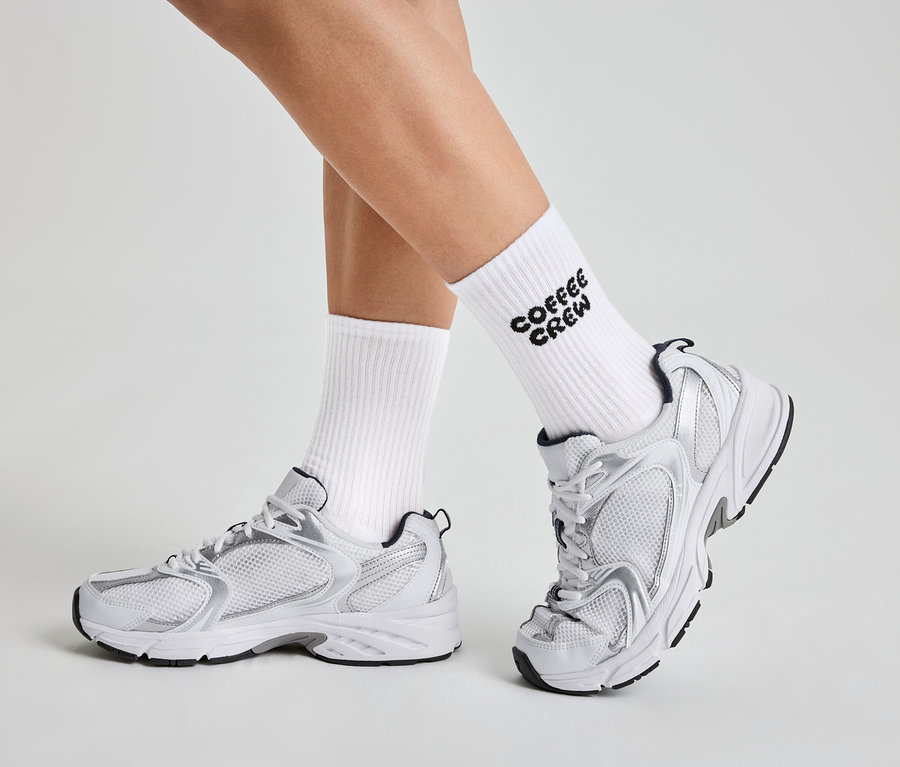 Beine einer Person in weißen Socken mit der Aufschrift "Coffee Crew" und weißen und silbernen Turnschuhen.
