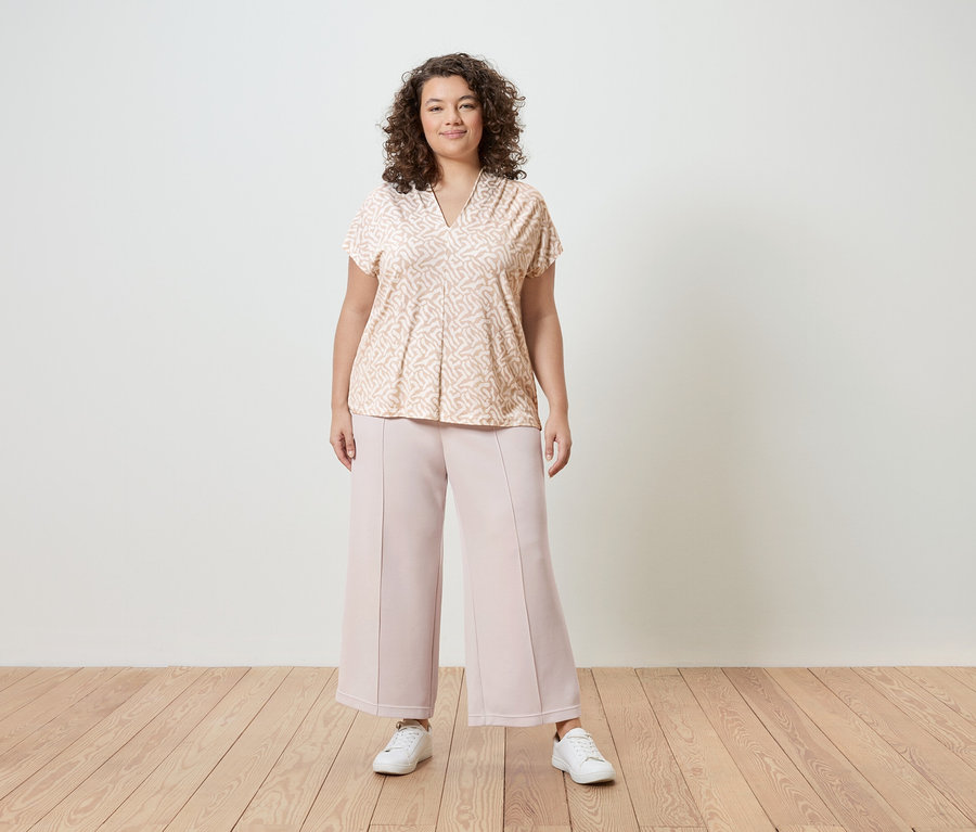 Eine Frau mit lockigem Haar trägt eine hellrosa gemusterte Bluse, rosafarbene Culottes und weiße Turnschuhe vor einer weißen Wand.