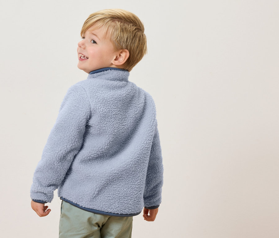 Ein kleiner Junge mit blonden Haaren trägt eine hellblaue Fleecejacke und hellgrüne Hosen, während er zur Seite schaut.