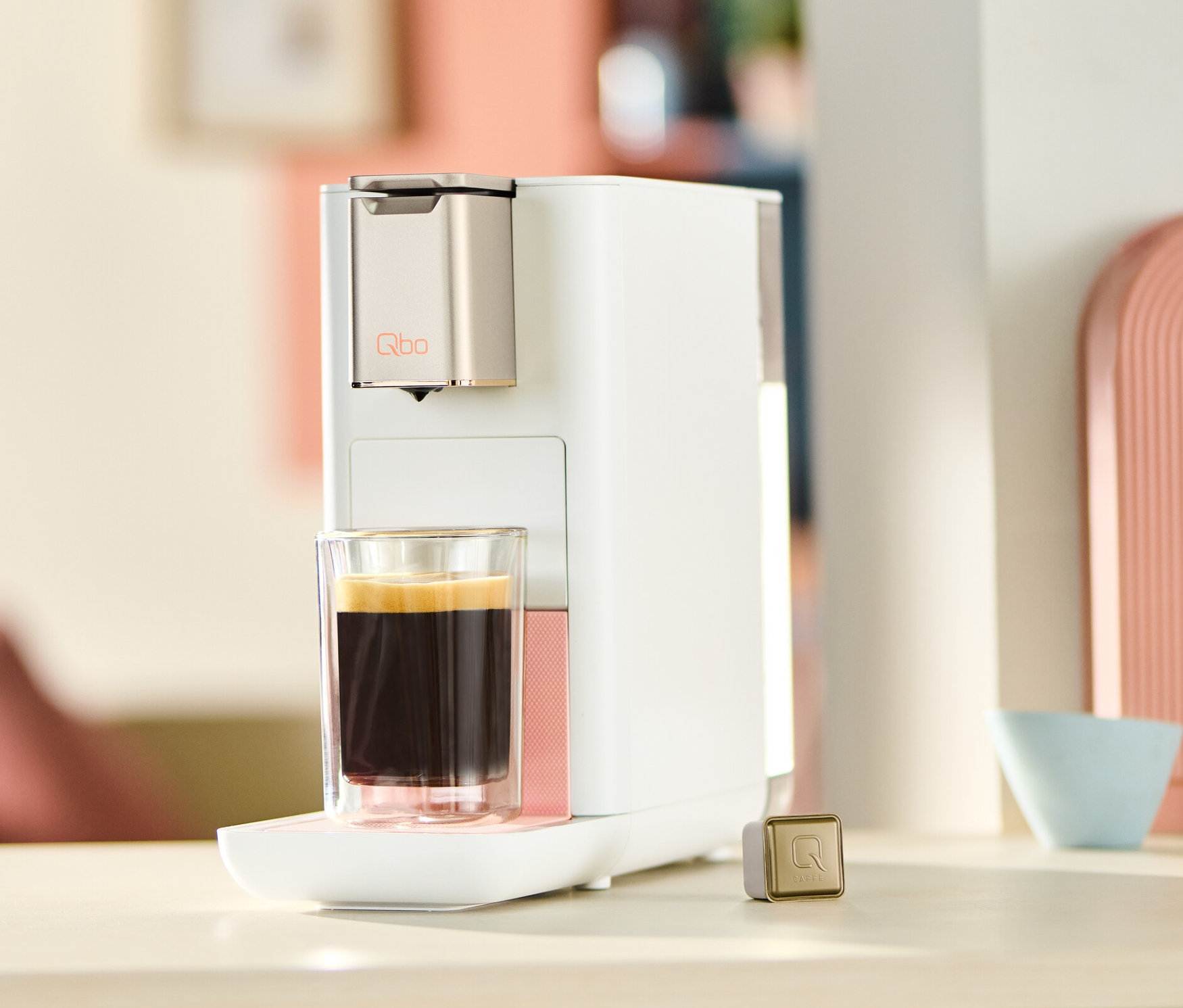 Qbo ICONIC x Pantone Special Edition Muted Clay Kaffeemaschine mit Tasse Kaffee und Kapsel auf dem Tisch.