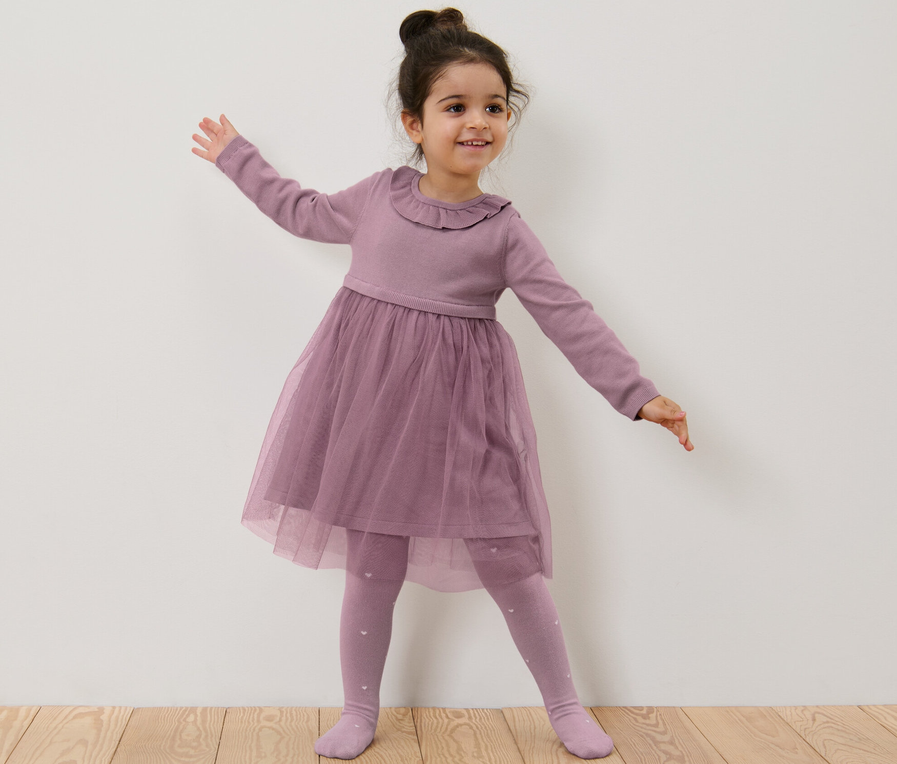 Lächelndes Mädchen im Kinder-Strickkleid mit Tüllrock steht vor einer weißen Wand.