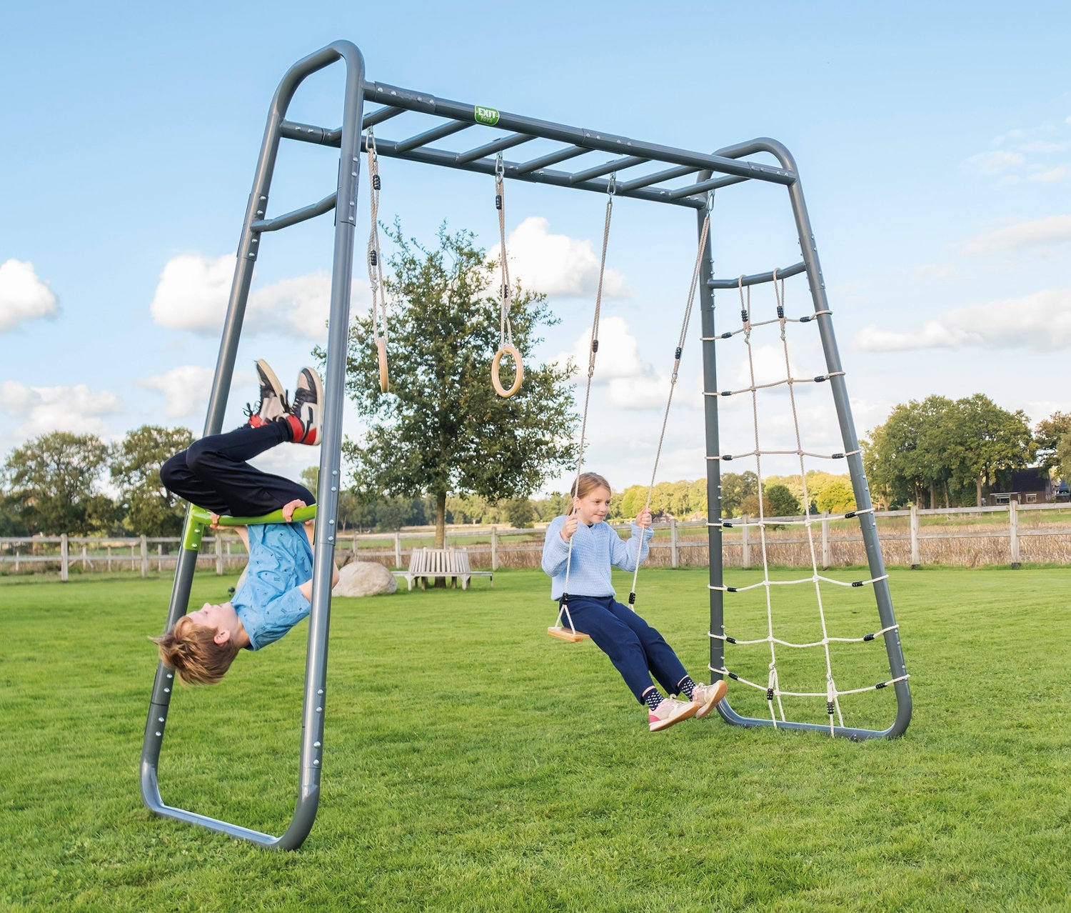 Ein Junge hängt kopfüber am EXIT Klettergerüst MB210, während ein Mädchen auf einer Schaukel sitzt.