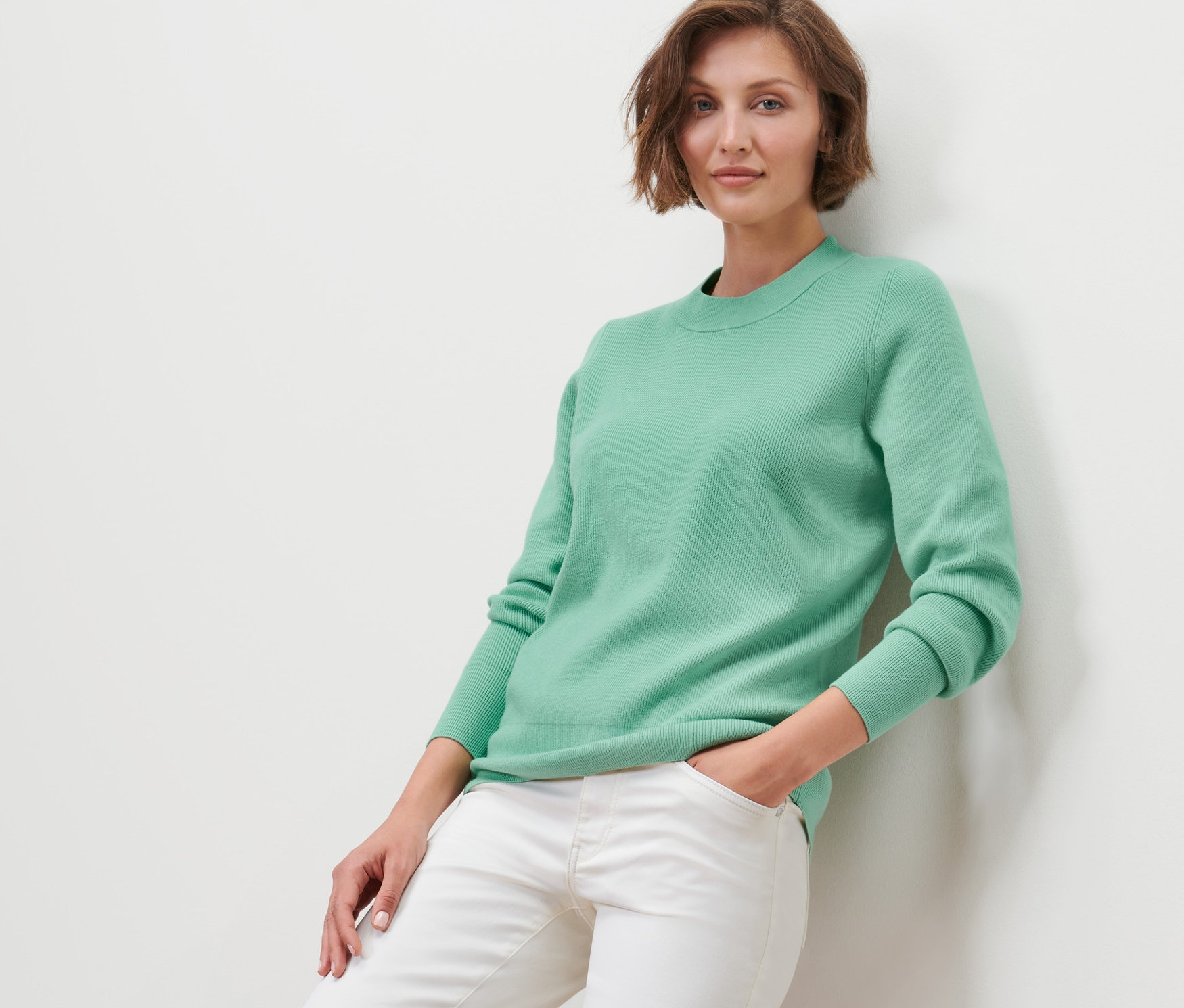 Frau lehnt an Wand, trägt Pullover und weiße Jeans.