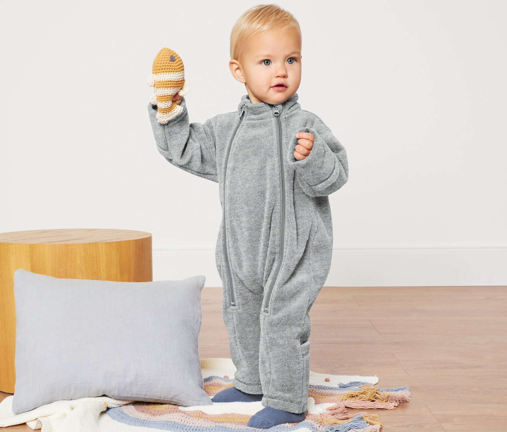 Kleinkind hält gehäkelten Fisch und trägt einen grauen Baby-Fleece-Einteiler.