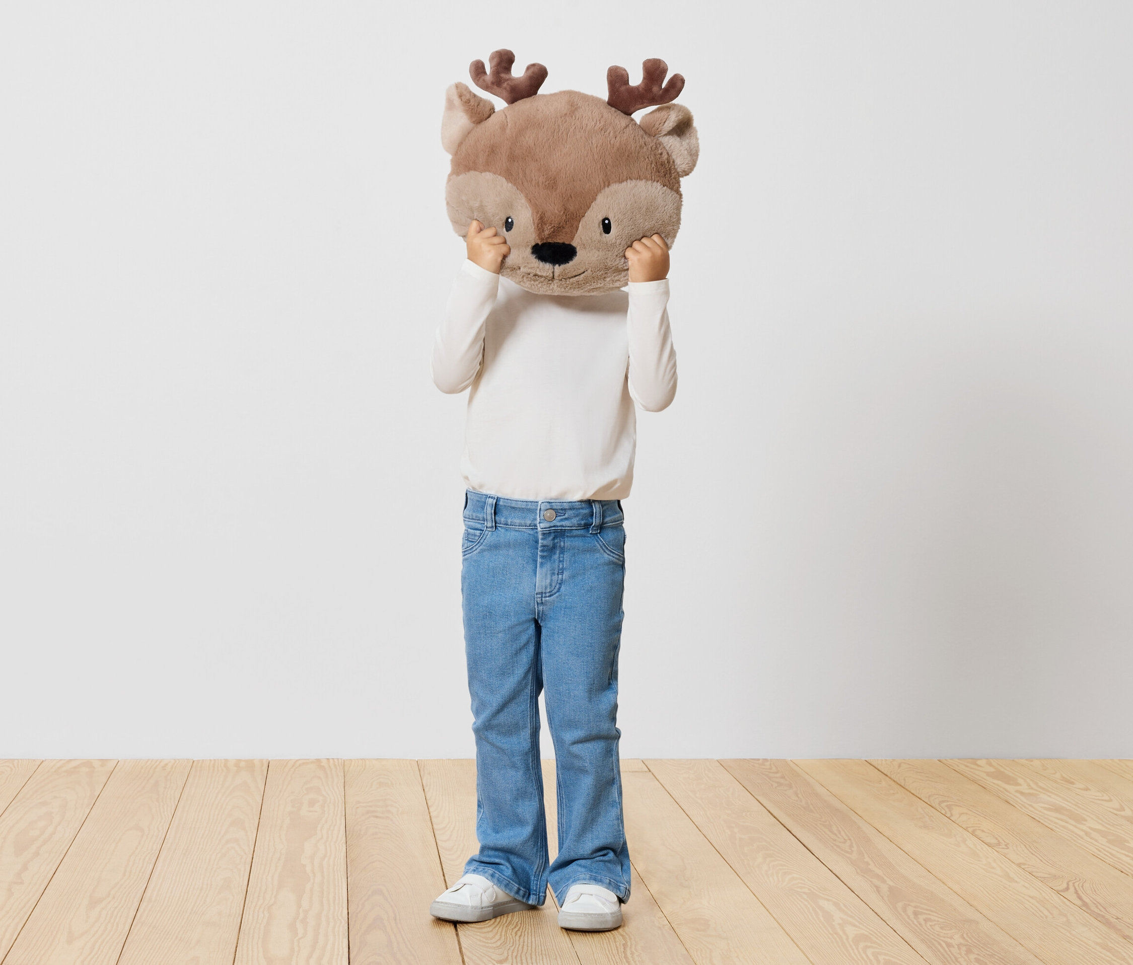 Ein Kind hält ein Stofftier-Rentier vor sein Gesicht, steht auf einem Holzboden vor einer weißen Wand.