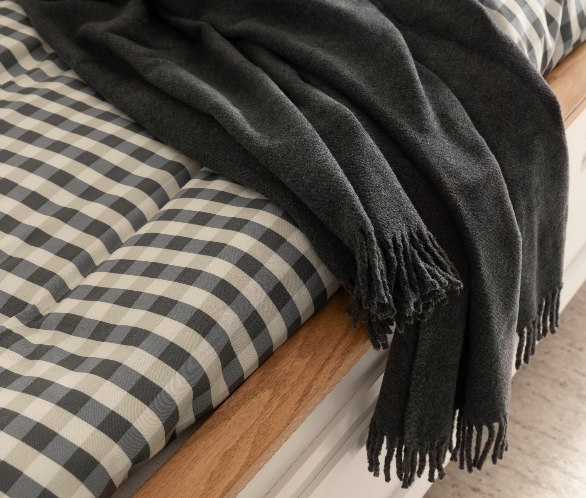 Eine graue Decke mit Fransen liegt auf einem Bett mit grau-weiß karierter Tagesdecke.