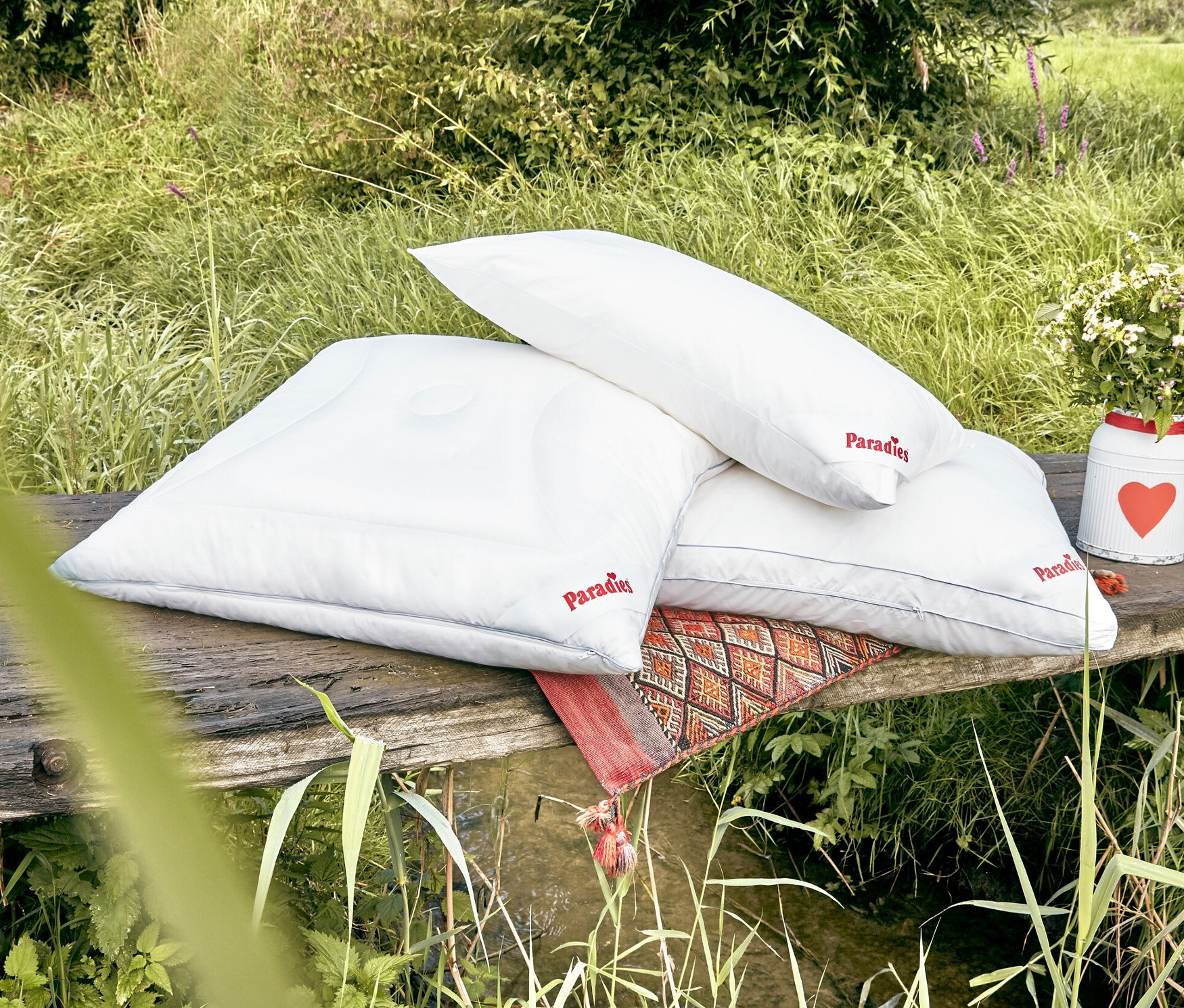 Drei weiße HanseTraum Kopfkissen liegen auf einem Holzbalken an einem Bach.