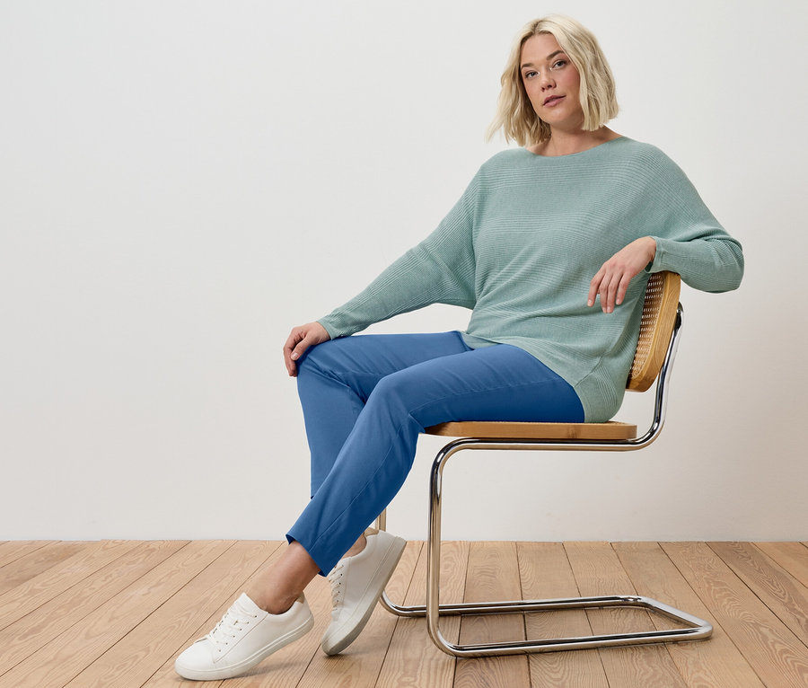 Blonde Frau sitzt auf einem Stuhl, trägt eine blaue Stretchhose und einen salbeifarbenen Feinstrickpullover.