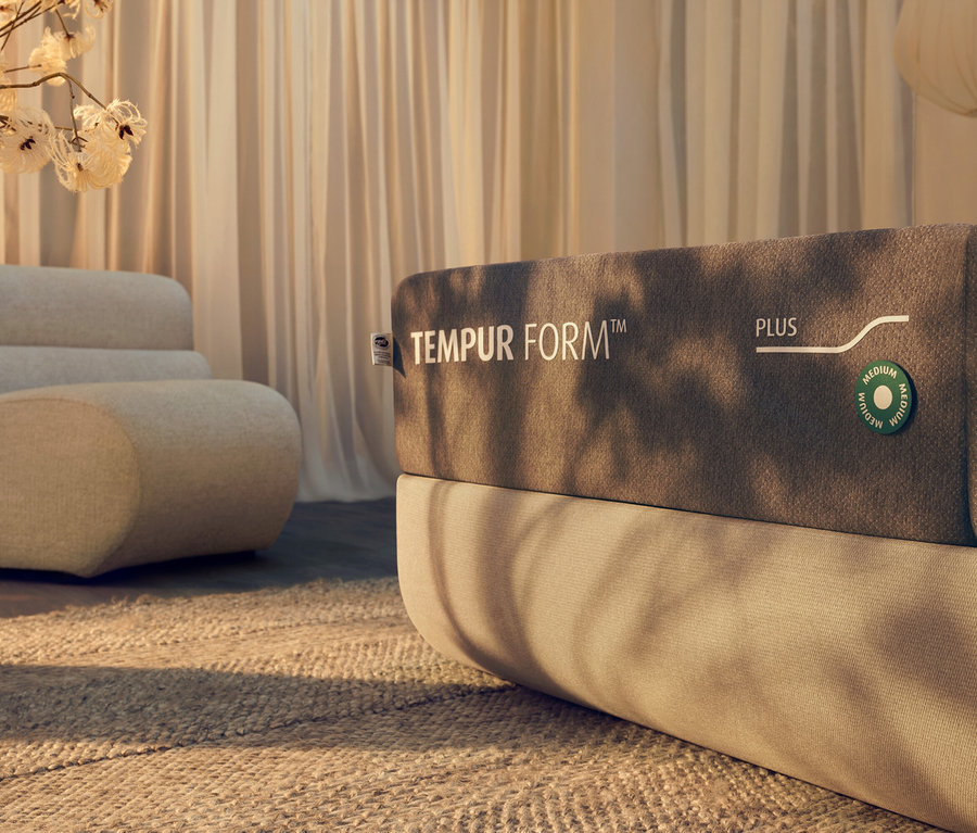 Nahaufnahme einer TEMPUR Form™ Plus Matratze medium auf einem Bett mit beigem Kopfteil, im Hintergrund ein beiger Sessel.